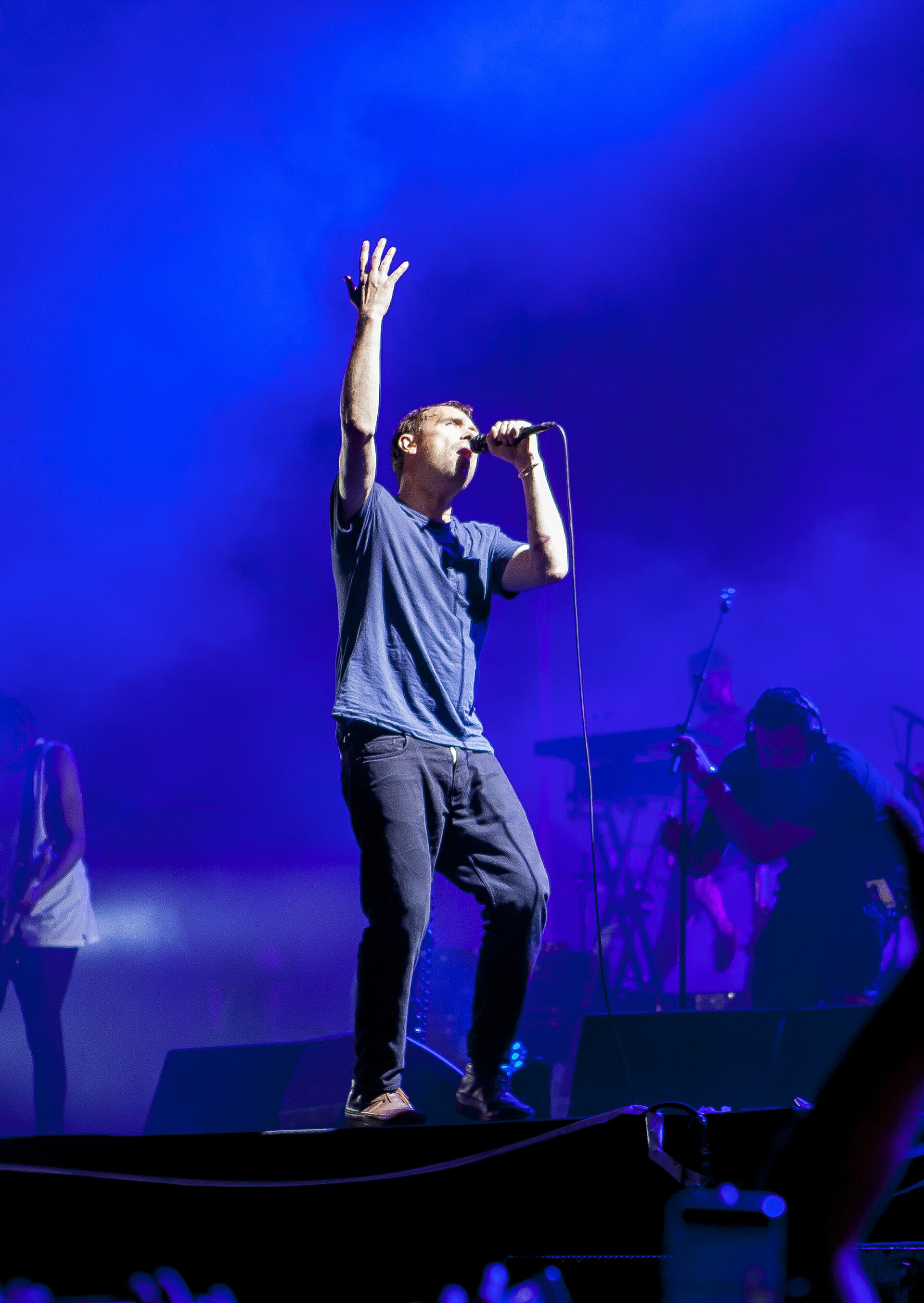 A singer in a blue t-shirt stands at the front of a stage with one arm raised high, microphone at his mouth, his band lit deep blue behind him
