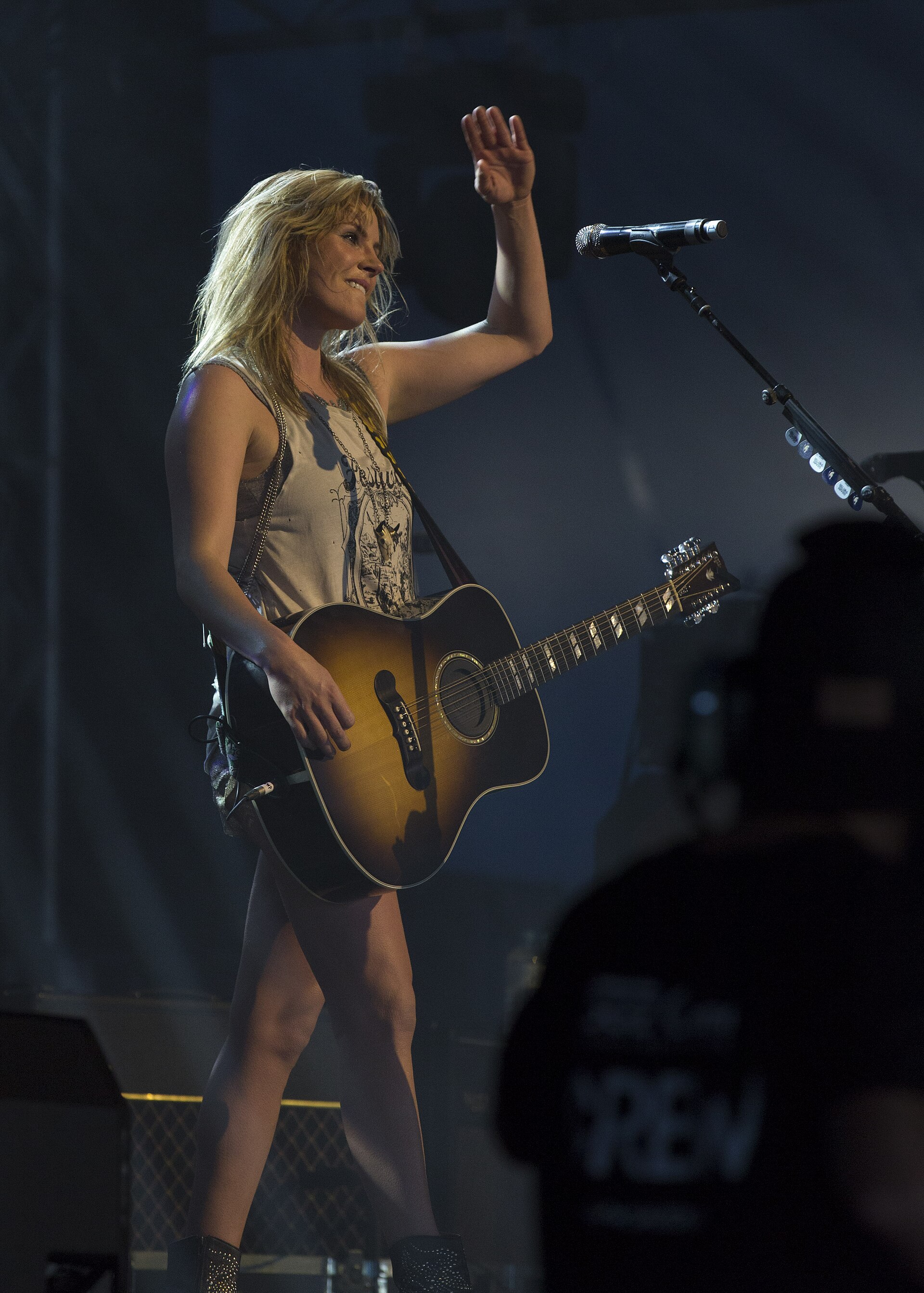 Grace Potter raising one hand to the crowd on a dark stage with an acoustic guitar slung across her, a microphone on a boom stand beside her