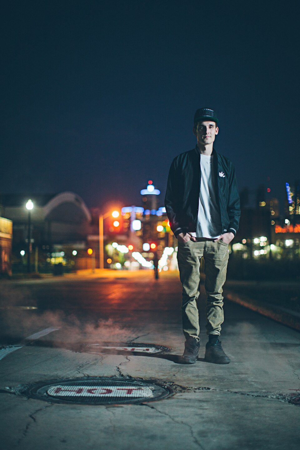 GRiZ stands on a city street at night in a cap and a bomber jacket, hands in his pockets, steam rising from a manhole behind him.