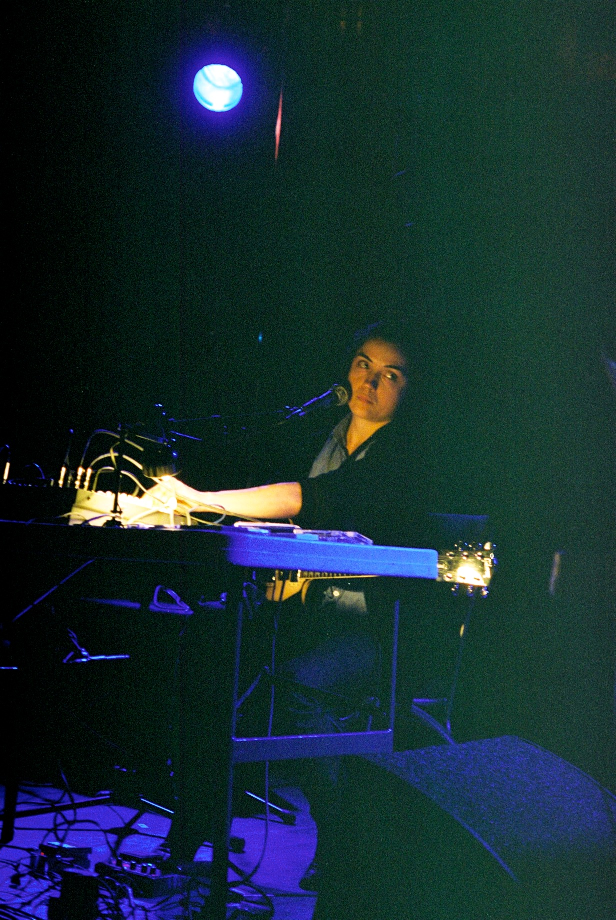 Liz Harris sits behind a table of pedals and a lap steel guitar, lit blue, singing into a microphone.
