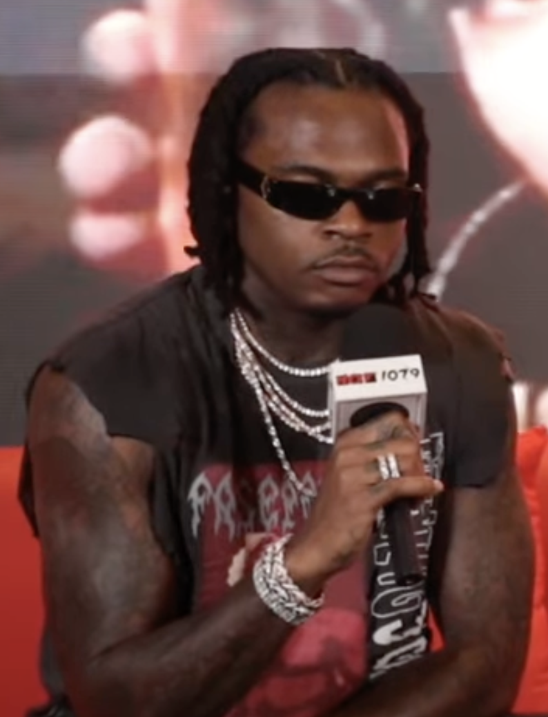 A rapper in dark sunglasses, a sleeveless black tee and silver chains holds a branded radio microphone during an interview