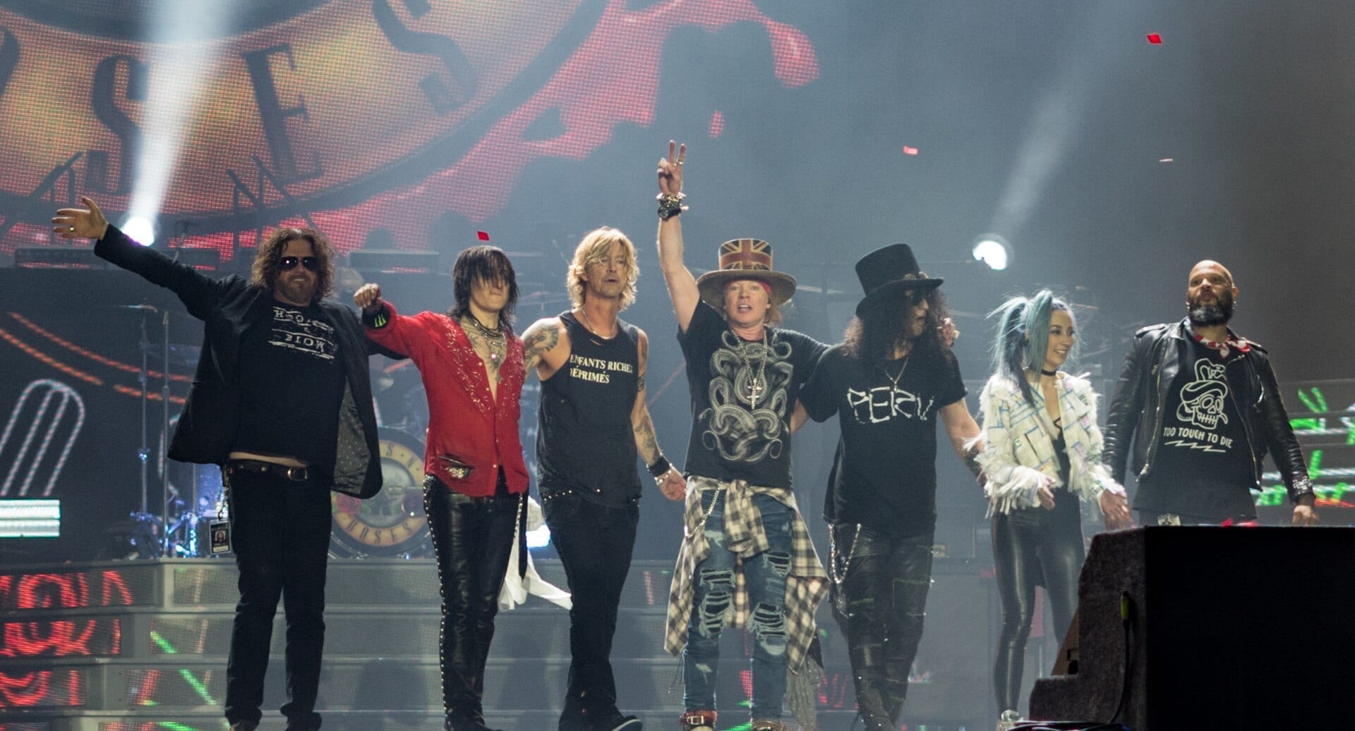 Guns N' Roses take a bow together on stage in a line, including Axl Rose in a top hat, Slash in a top hat with curly hair, and Duff McKagan.