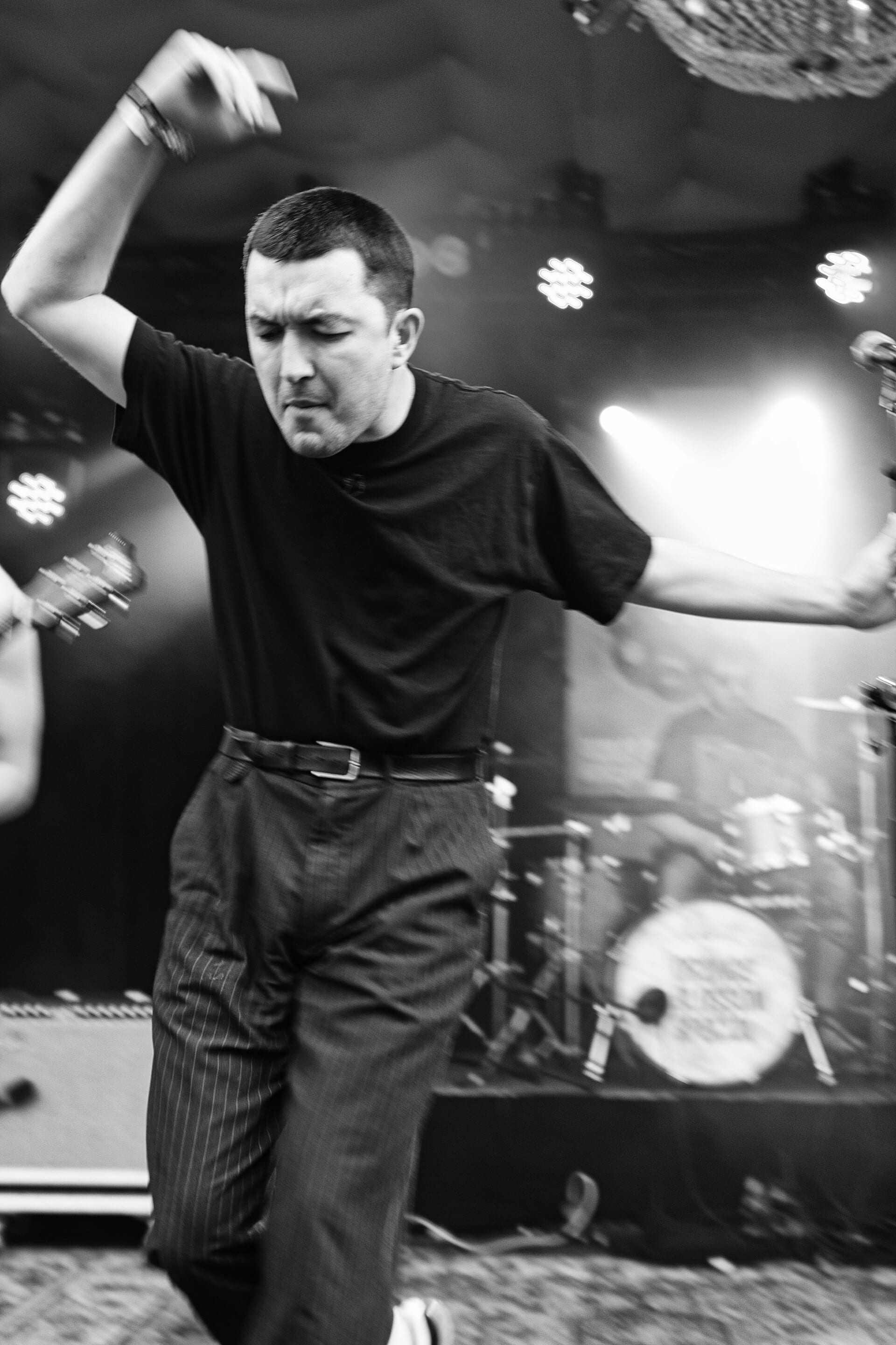 A black-and-white live photo of a Gurriers band member mid-movement on stage, arm raised, wearing a black T-shirt and pinstriped trousers, with a drum kit and stage lights visible behind him.