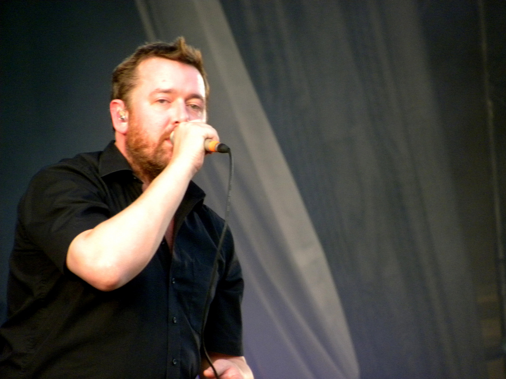 Guy Garvey sings into a handheld microphone in a black short-sleeved shirt, an in-ear monitor visible.