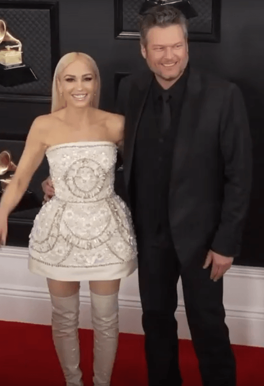 Gwen Stefani, in a white beaded strapless dress and matching thigh-high boots, stands arm in arm with Blake Shelton in a black suit on a red carpet.