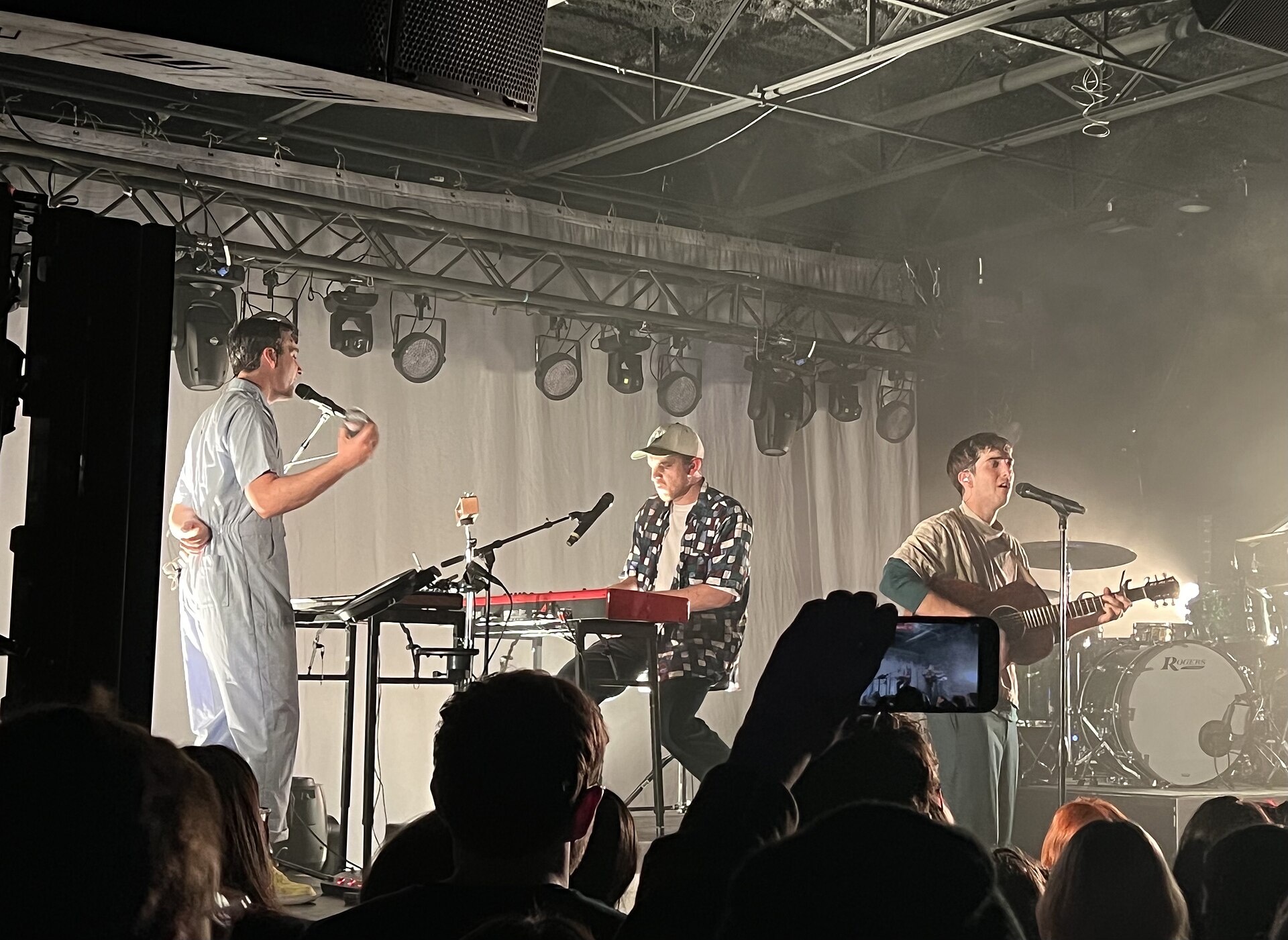 Three members of half-alive on a small club stage, one singing at a mic stand, one at a red keyboard and one playing an acoustic guitar.