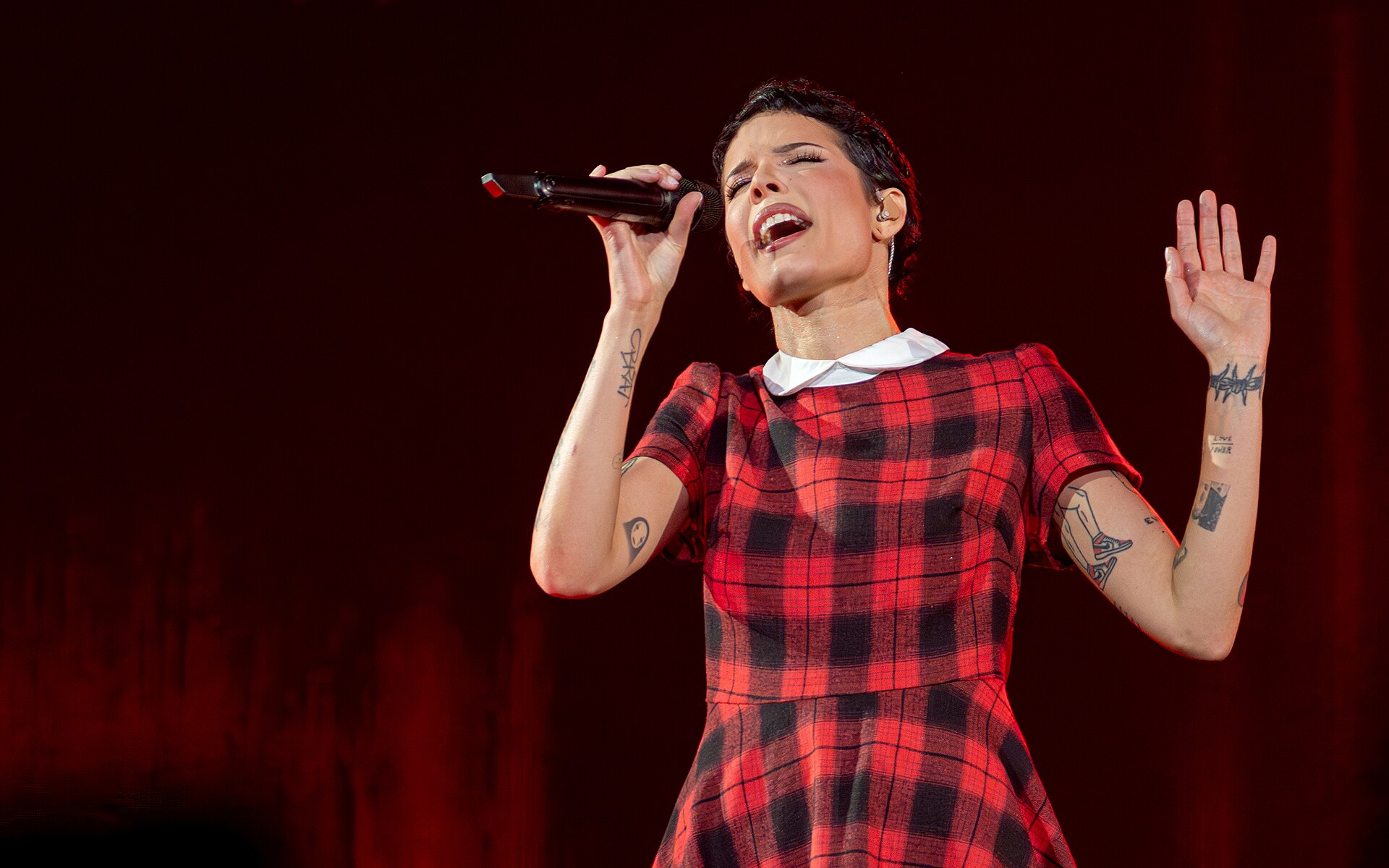 Halsey sings into a handheld microphone on a dark stage in a red tartan dress, short dark hair pushed back, tattooed arms bare.