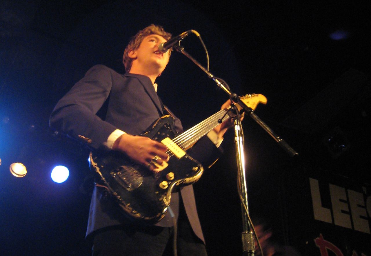 Hamilton Leithauser sings up into a microphone on a club stage, shot from below, playing a heavily worn black electric guitar in a navy suit jacket.