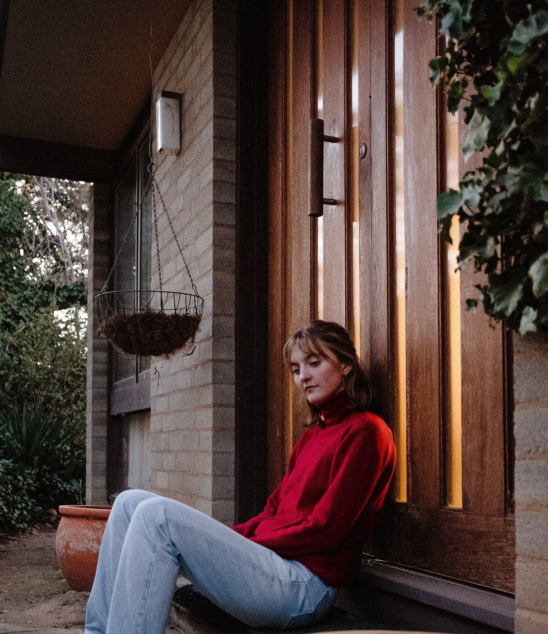 Hana Stretton sits on a porch step in a red turtleneck and jeans, leaning against a wooden door beside a hanging planter and ivy.