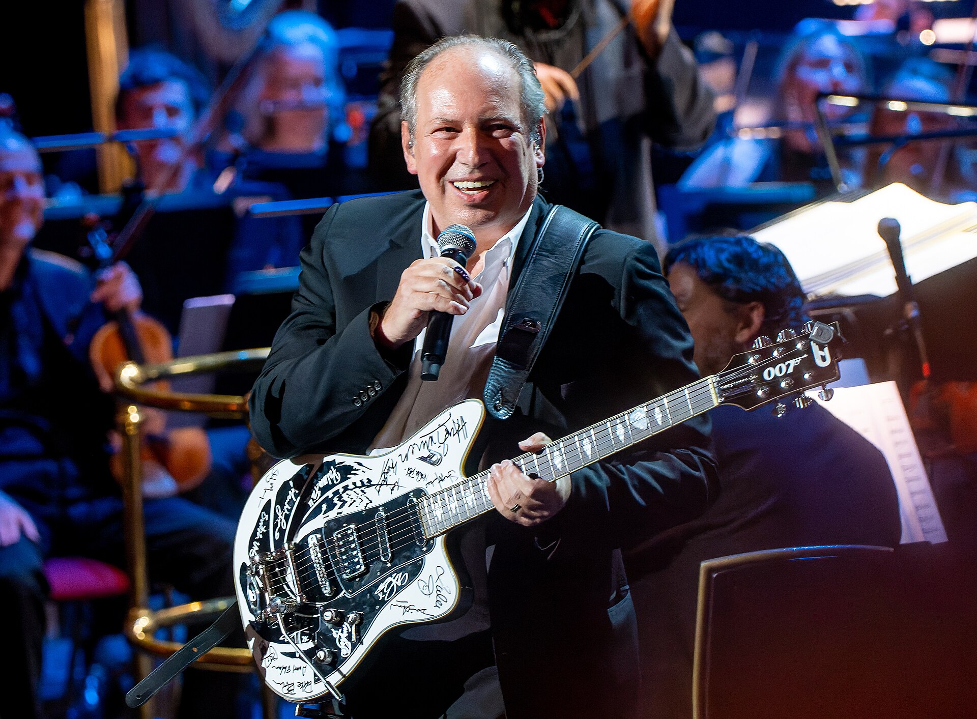 Hans Zimmer grins while speaking into a handheld microphone on stage, a signature-covered white electric guitar with a 007 headstock slung across his dark suit, an orchestra behind him.