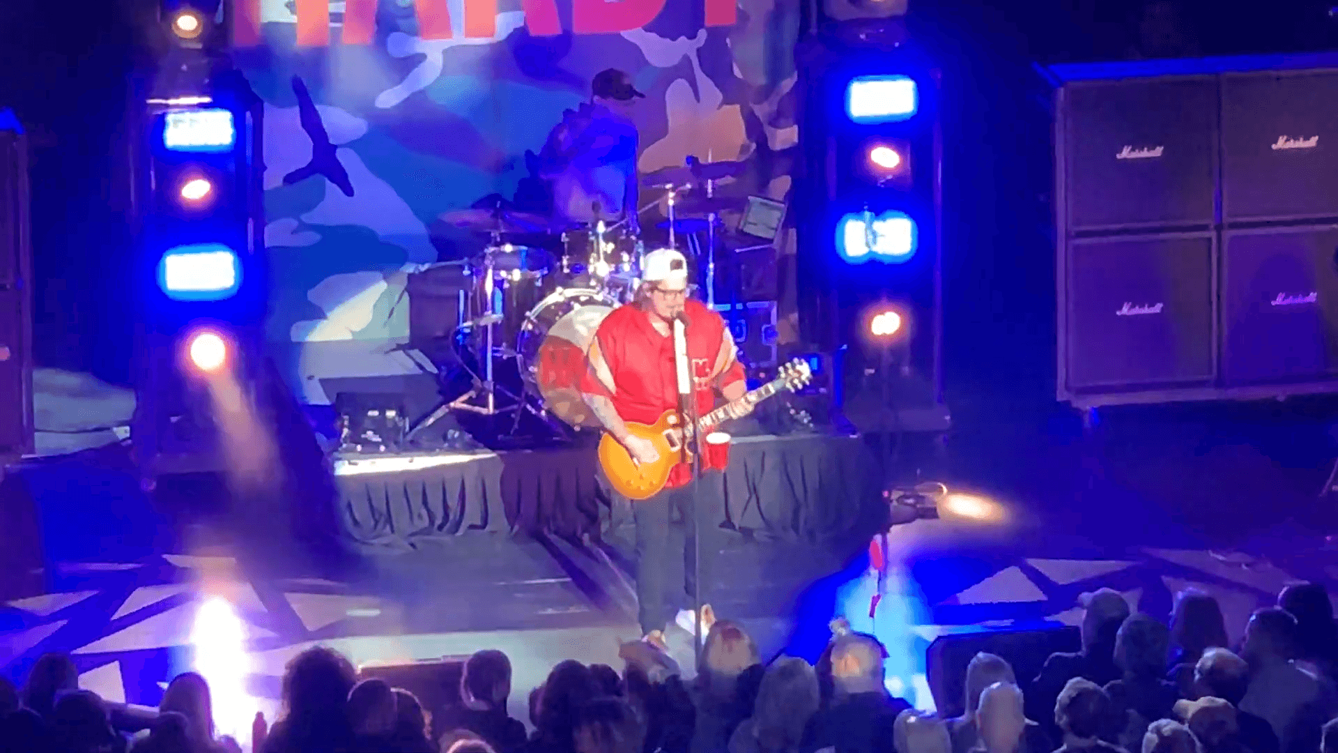 HARDY performs on stage in a backwards cap and red shirt, playing an electric guitar in front of a drum kit and amps, with his name lit up on the backdrop.