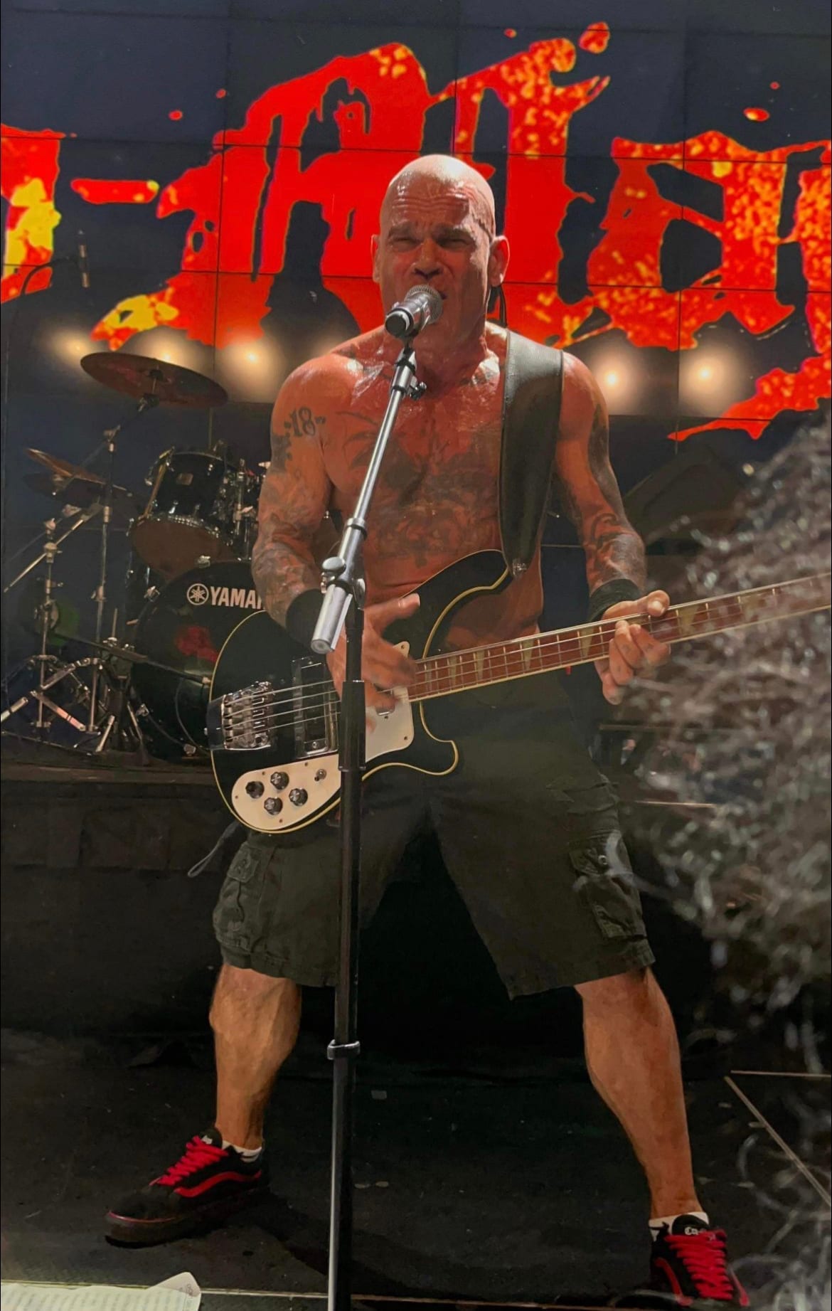 Harley Flanagan, shirtless and heavily tattooed, sings into a microphone while playing a black bass guitar on stage.
