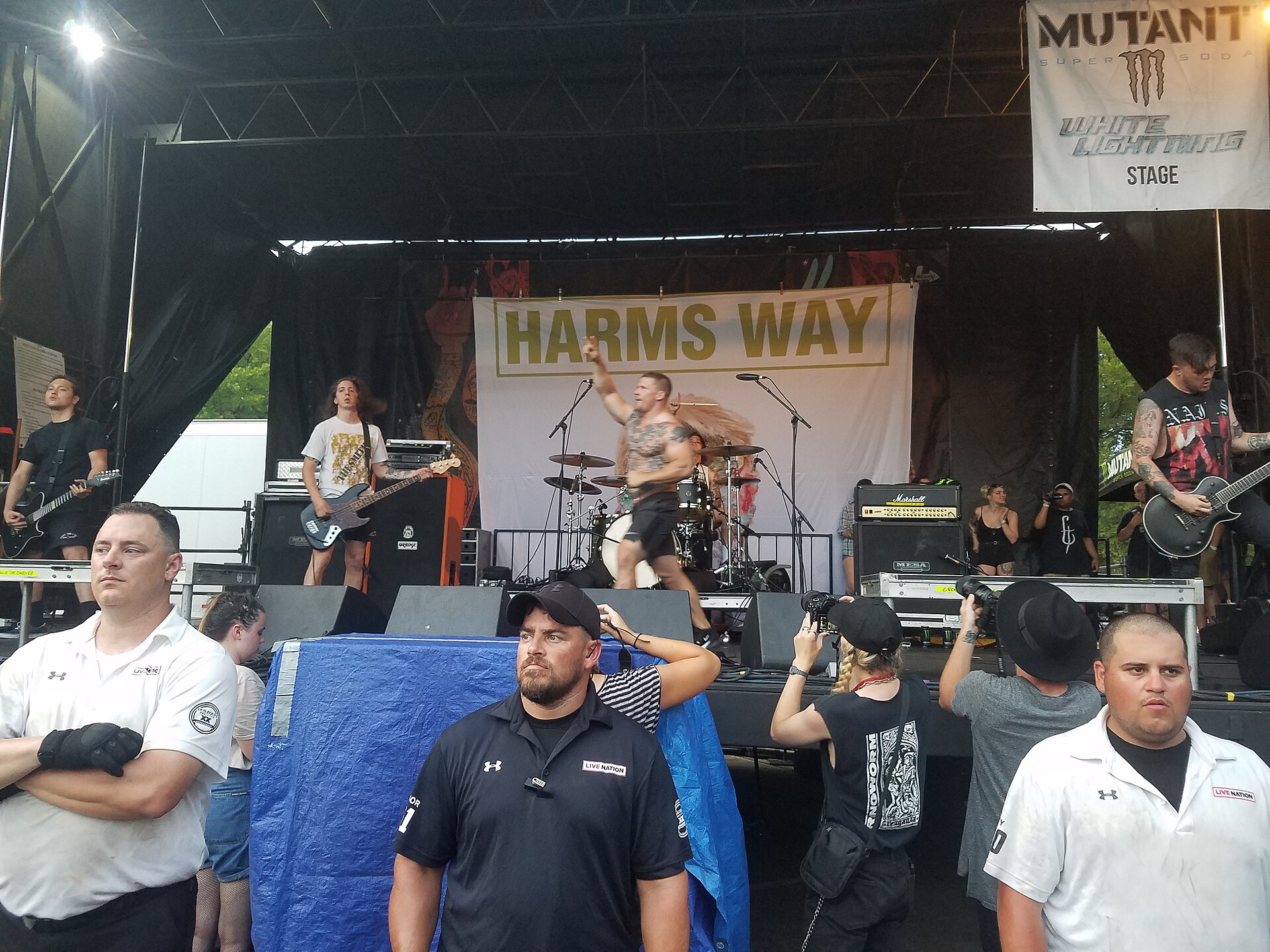 Harm's Way play a daylight stage in front of a large banner printed with the band's name, the singer crouched at the front with a microphone.