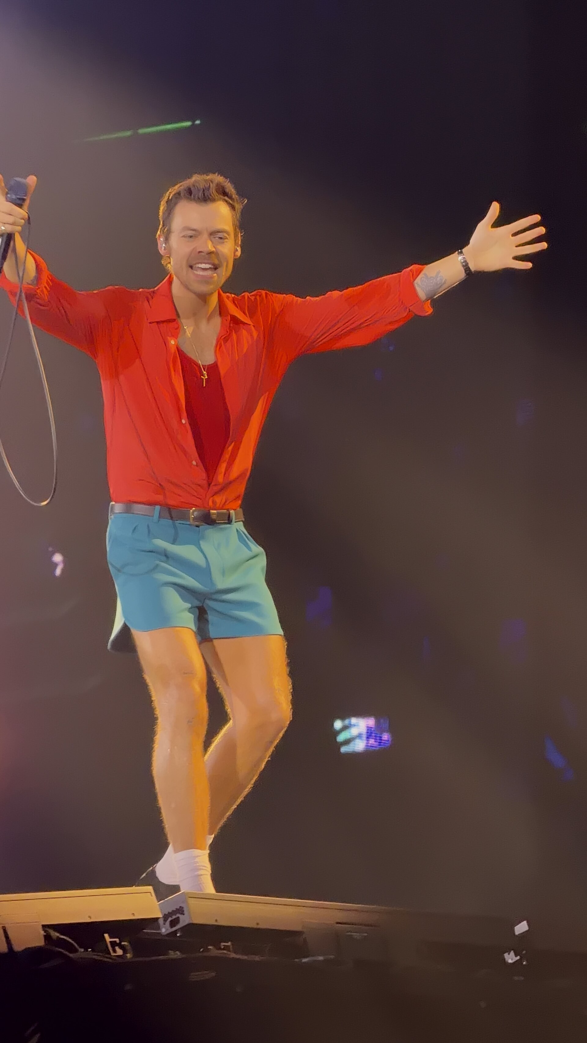 Harry Styles stands on a stage platform with his arms flung wide, in an open red shirt and teal shorts, a microphone in one hand.