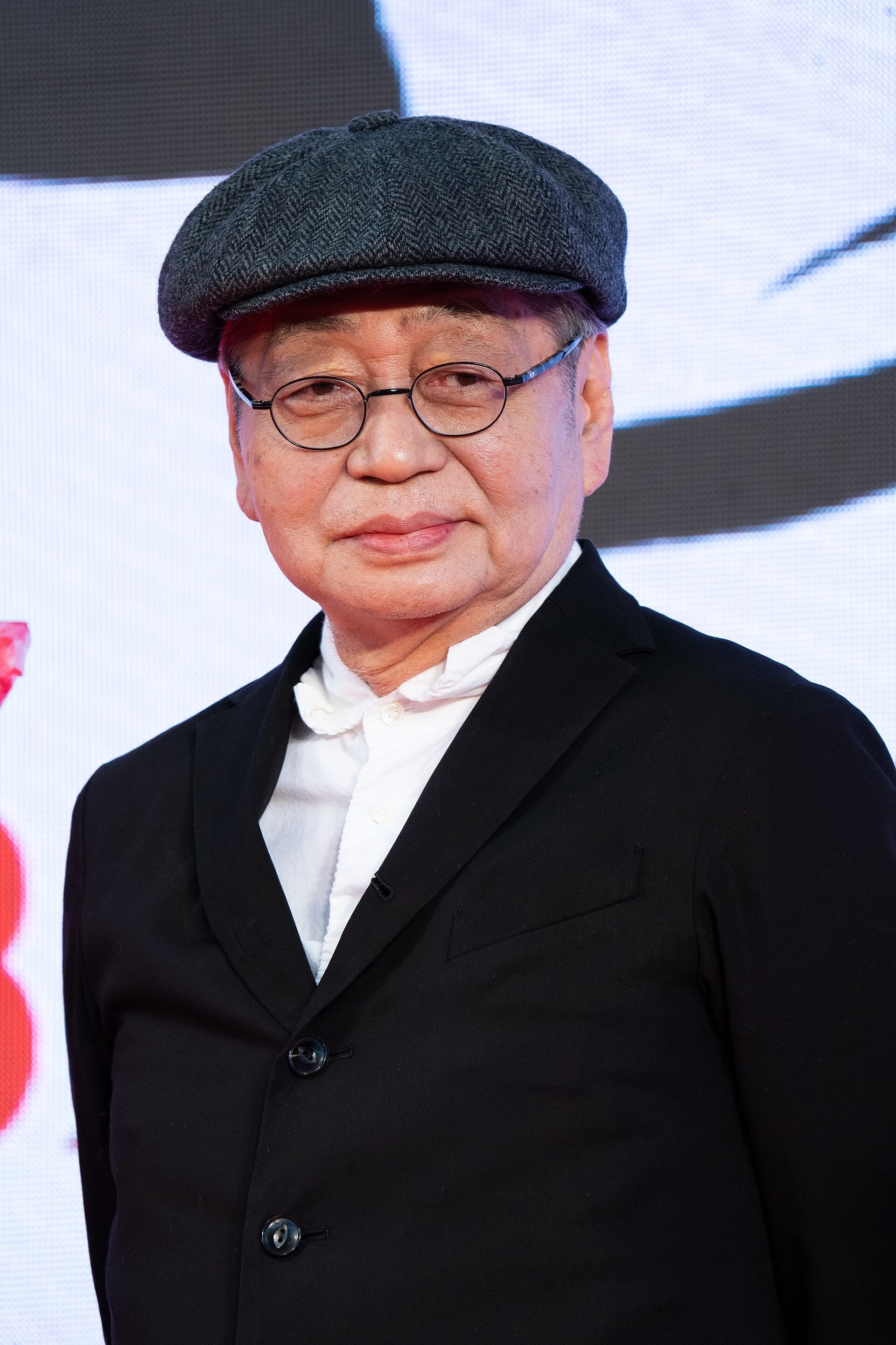 Haruomi Hosono, wearing round glasses and a newsboy cap, poses in a dark blazer at a film festival event.