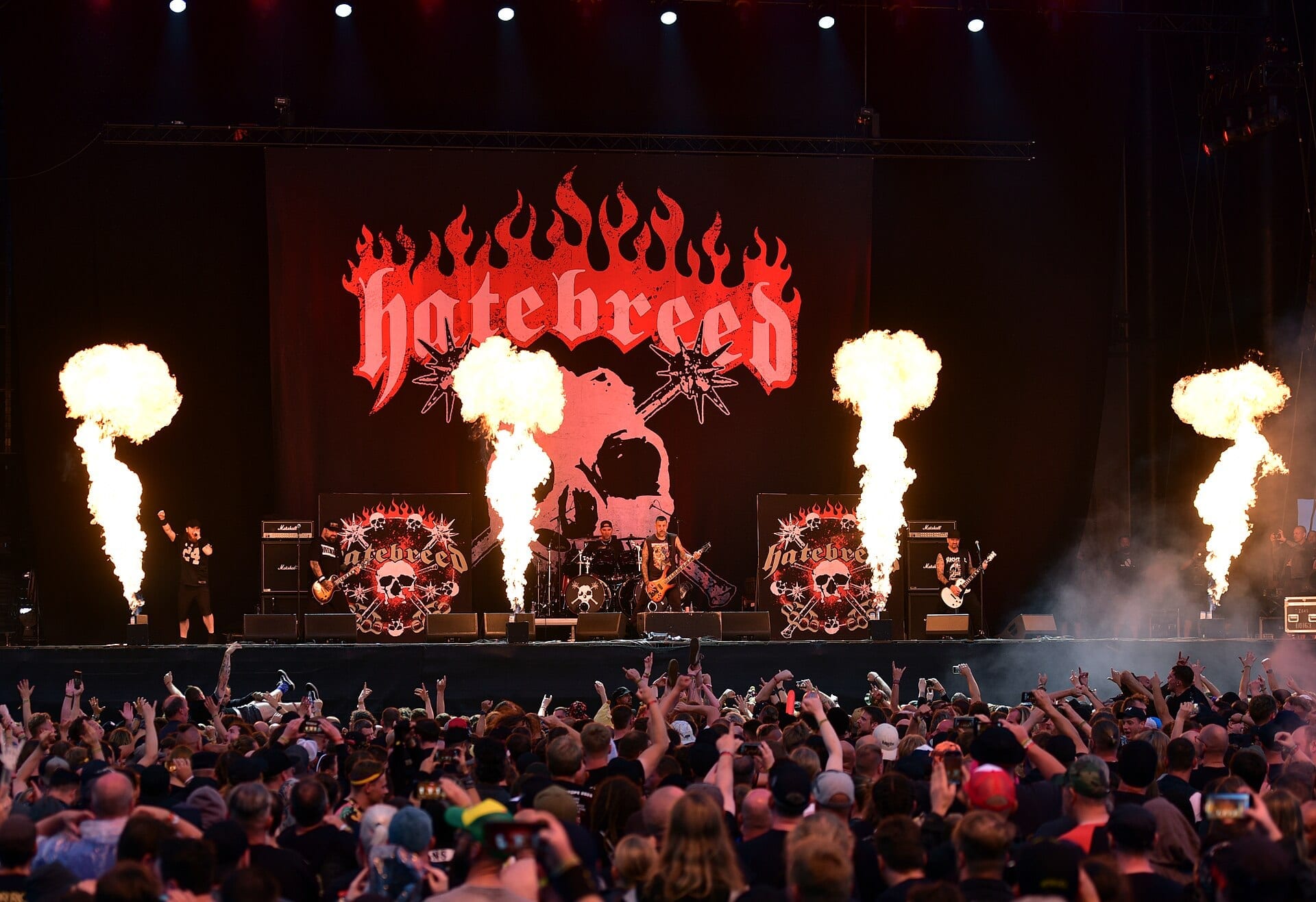 Hatebreed play a festival stage under a red flaming logo banner as four jets of fire shoot up across the front, a packed crowd with raised hands in the foreground.
