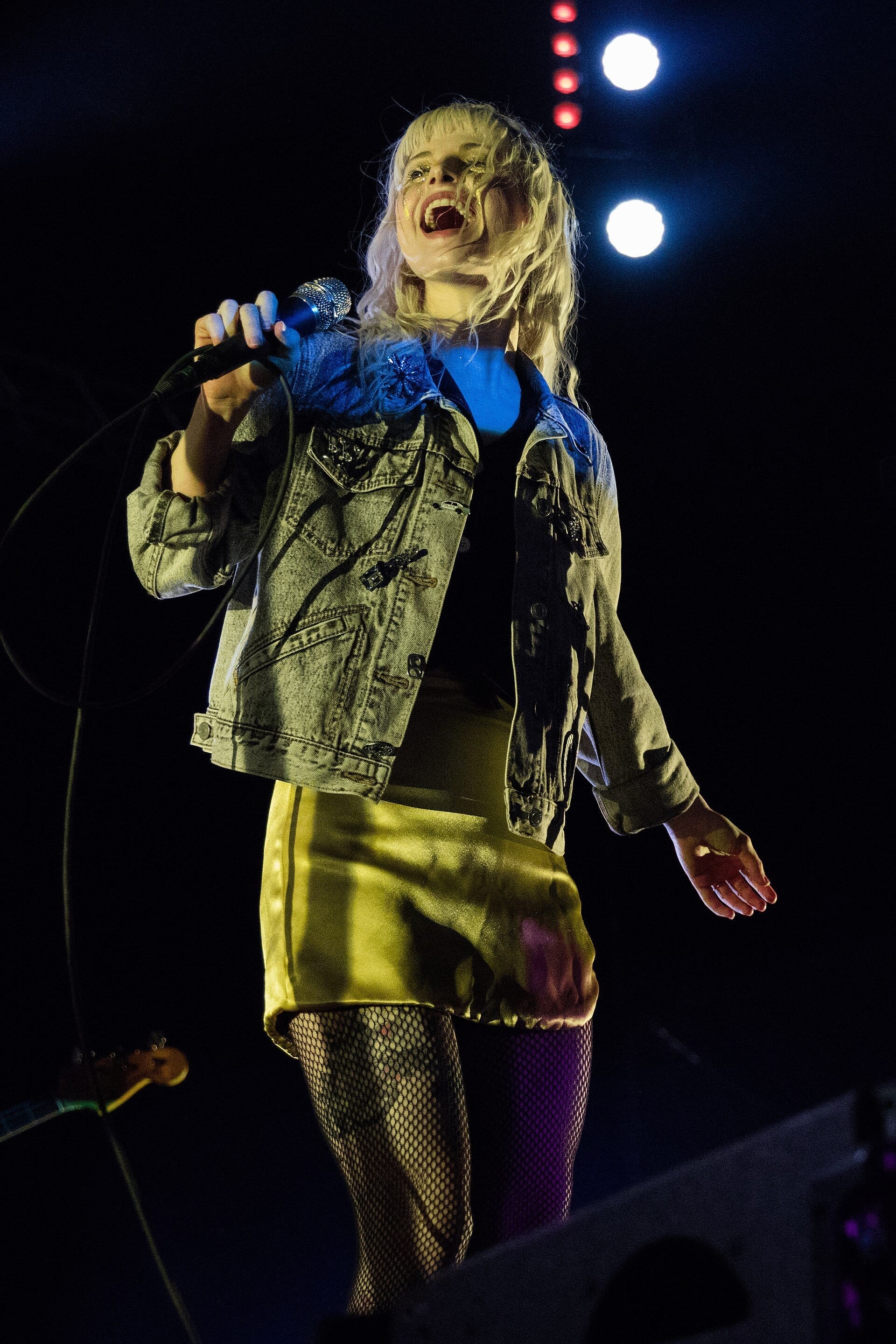 Hayley Williams, with pale blonde hair and a denim jacket over a satin dress, sings into a handheld microphone on a dark stage lit by two bright spotlights.
