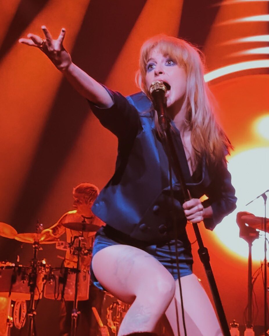 Hayley Williams sings into a mic on a stand with one arm flung out toward the crowd, lit orange, a drummer behind her.