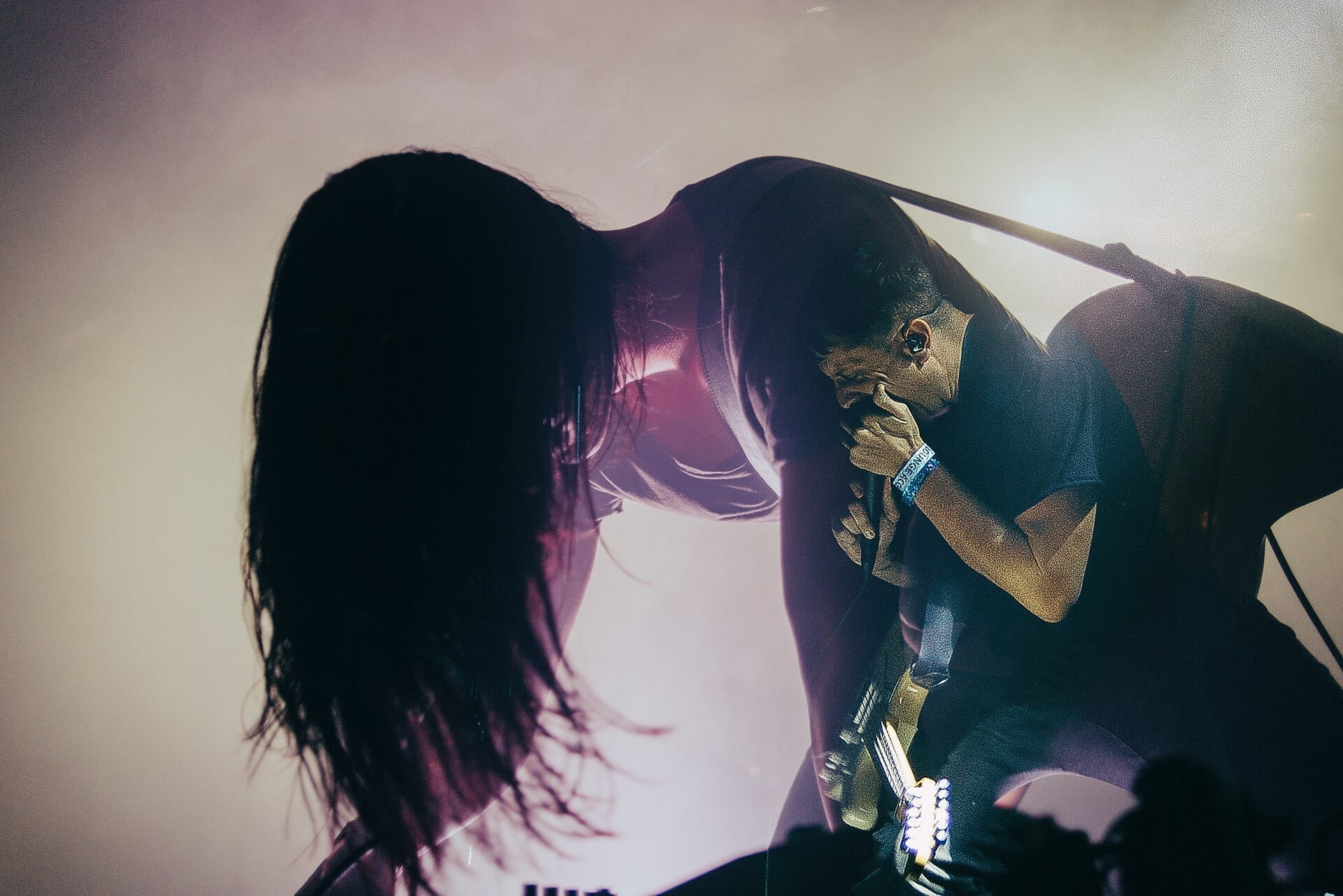 A member of the band HEALTH singing into a microphone on a foggy, backlit stage.