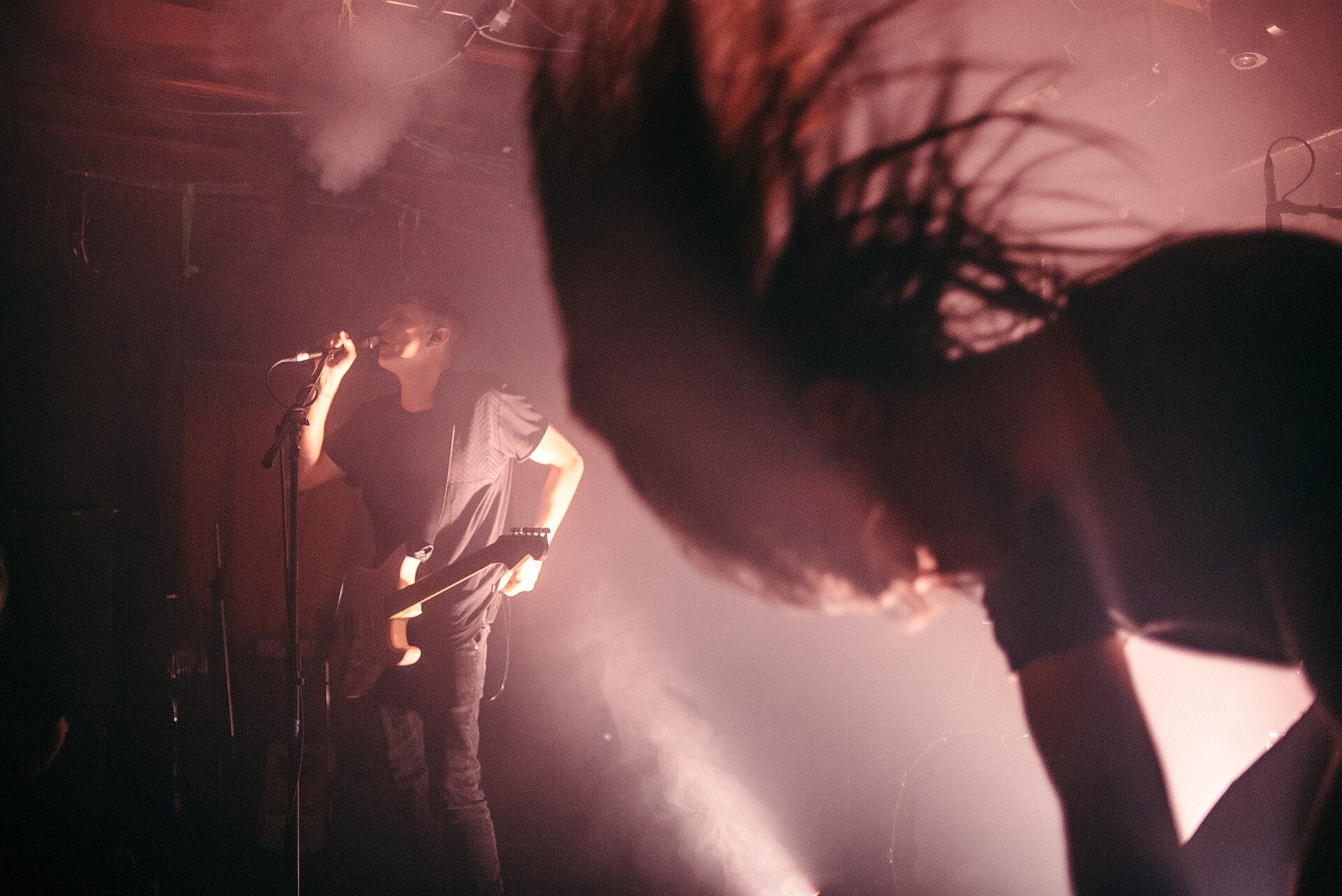 A HEALTH guitarist sings into a mic stand in red haze while a bandmate whips their long hair through the foreground.