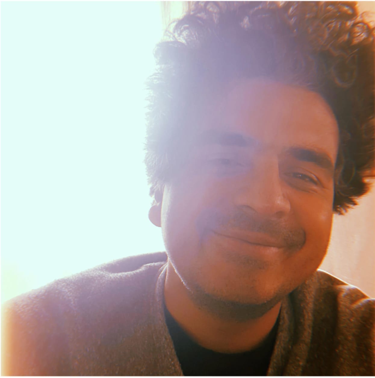 A sunlit close-up self-portrait of Helado Negro smiling, with curly hair backlit by warm light.