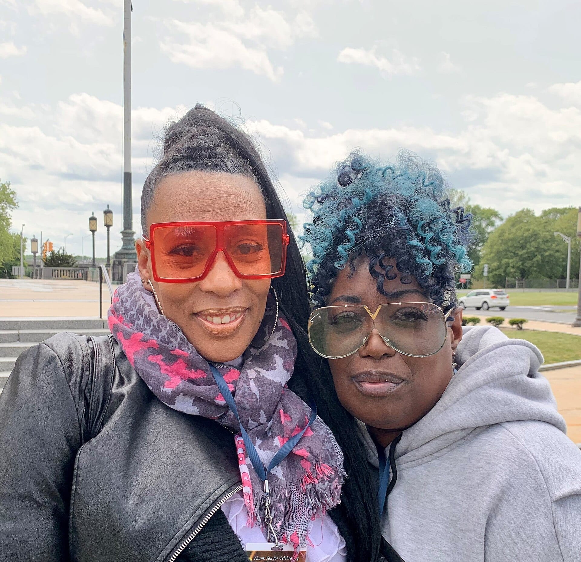 Two women pose together outdoors for a selfie, one wearing red sunglasses and a patterned scarf, the other with teal-tinted curly hair and round tinted sunglasses, in front of a plaza with lamp posts and trees.