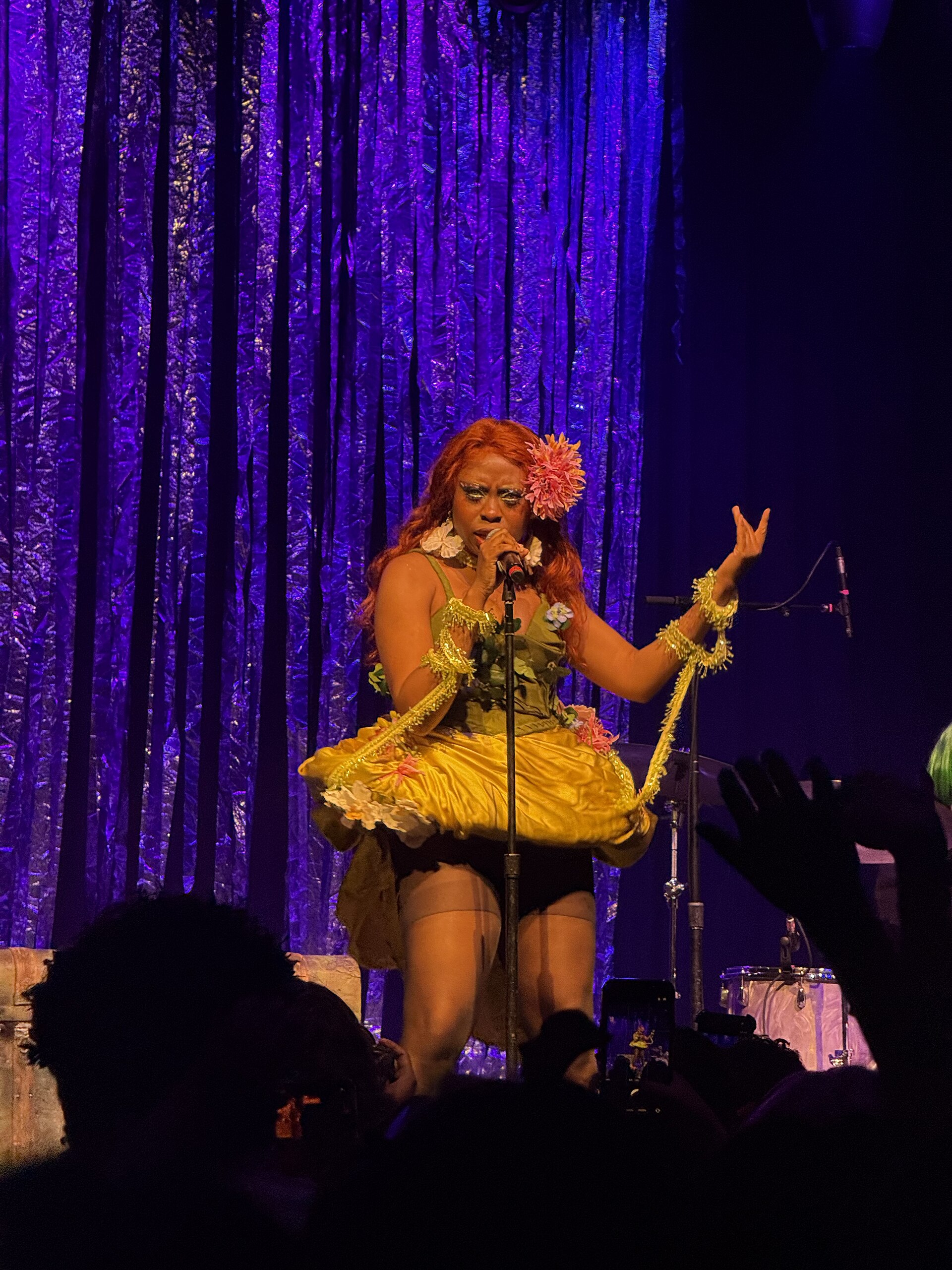 hemlocke springs sings into a microphone on stage in a yellow and green flower-trimmed costume, long orange hair with a bloom pinned in it, one hand raised, against a purple foil curtain.