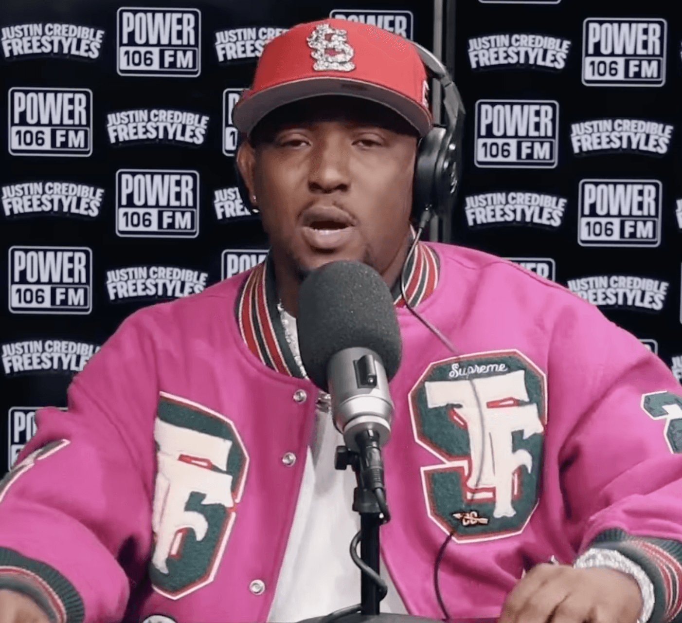 Hit-Boy raps into a microphone wearing headphones, a red fitted cap and a pink varsity jacket, in front of a Power 106 FM step-and-repeat backdrop.