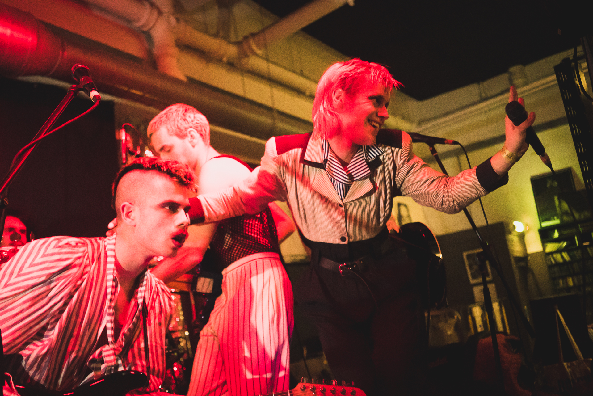 Three members of HMLTD crowd together on a low red-lit stage in striped shirts and pinstriped trousers, one reaching for a microphone.