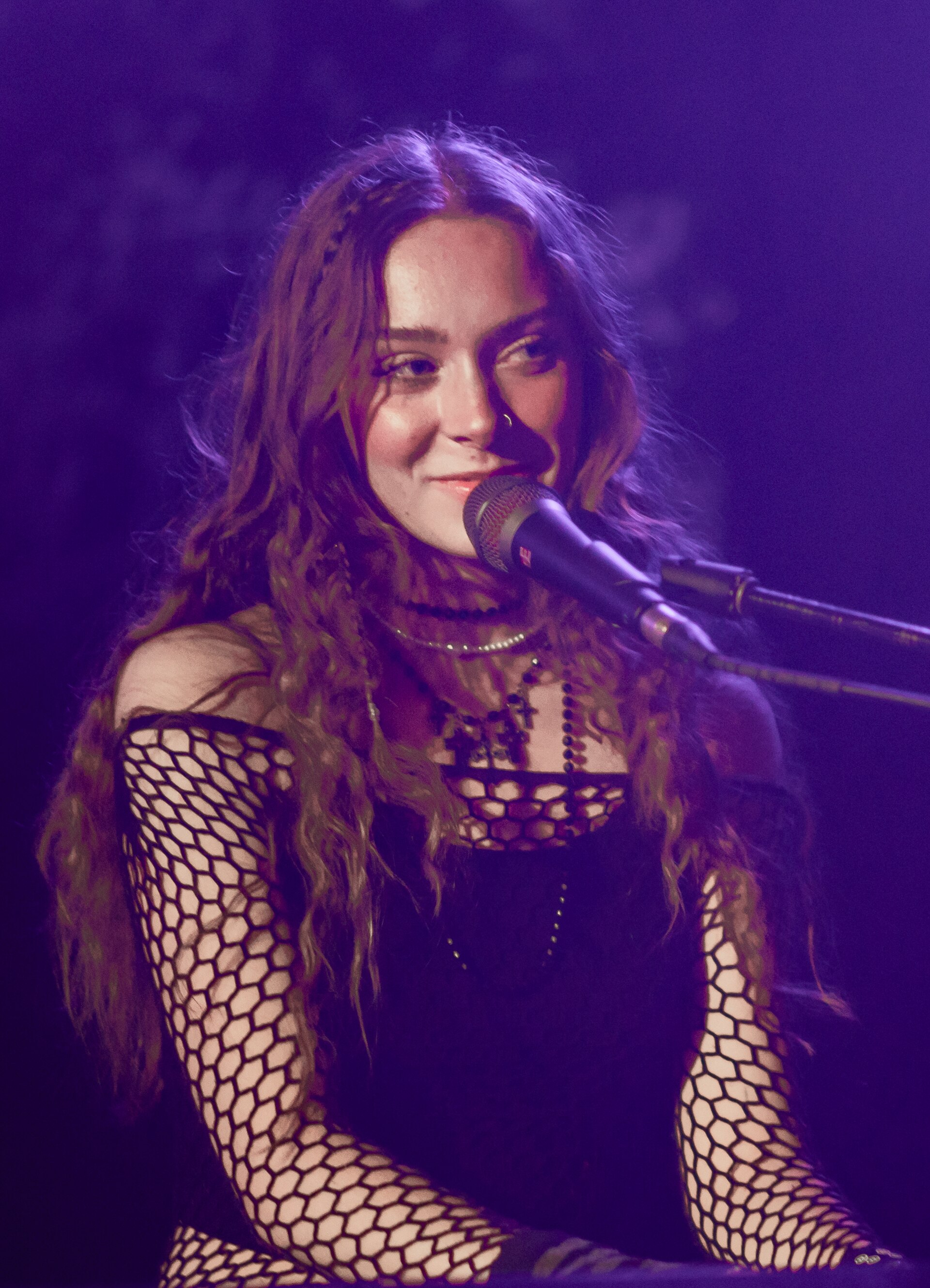 Holly Humberstone at a microphone under purple stage light, long wavy hair loose, wearing a black fishnet top over a black dress and layered necklaces