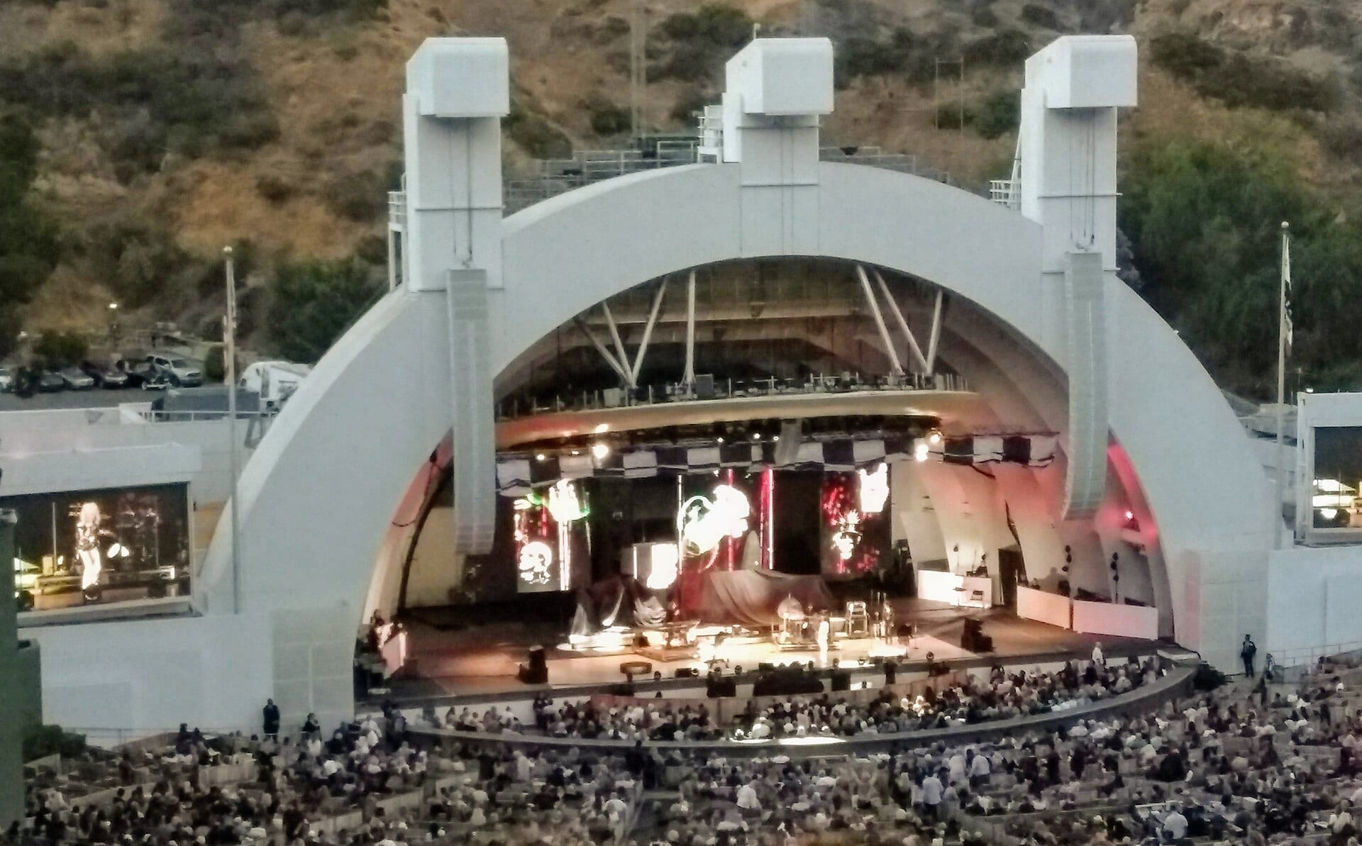 The Hollywood Bowl amphitheater, its concentric white concert bands lit at dusk, with a band performing on stage before a full crowd.