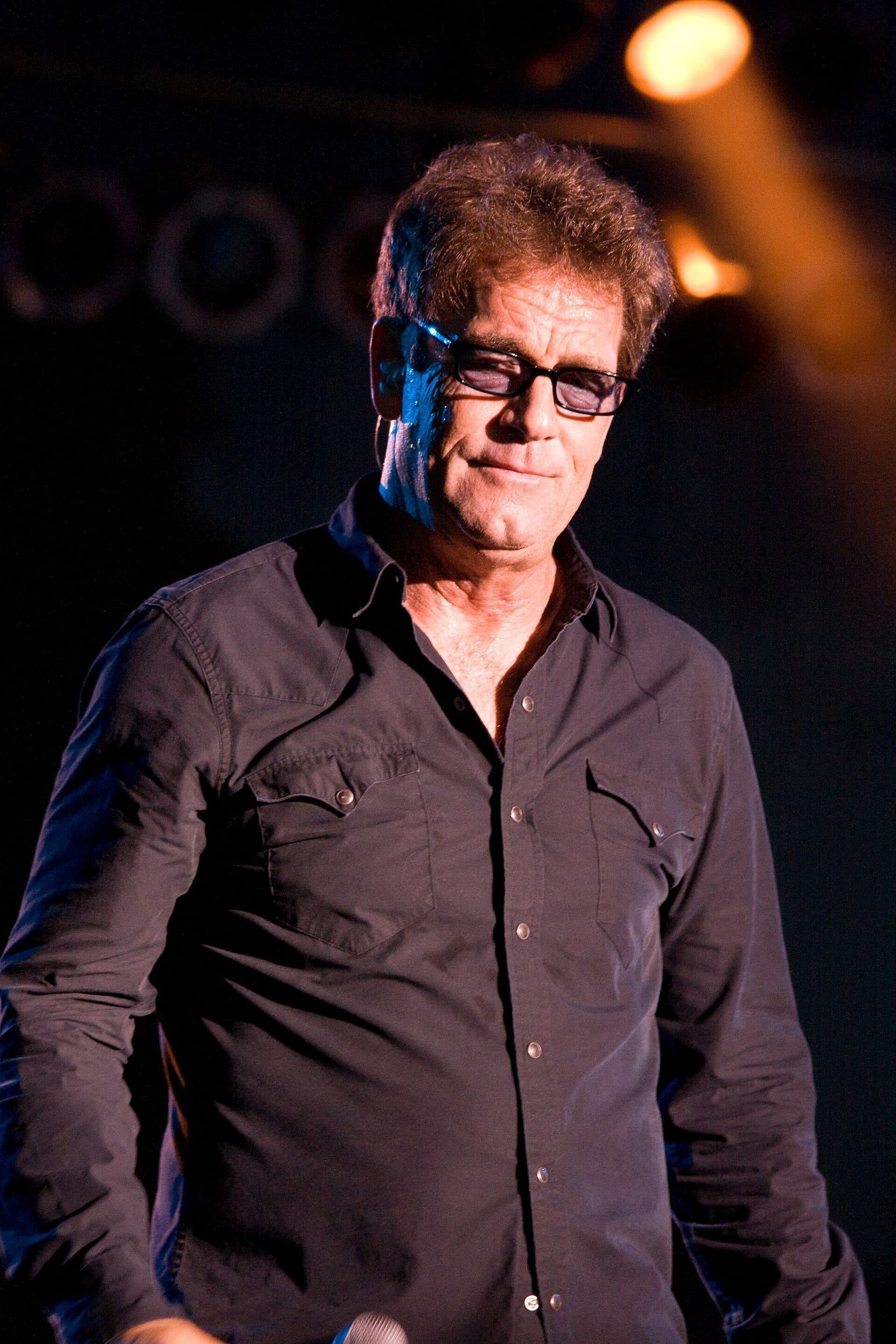 Huey Lewis stands in a dark shirt and rectangular glasses on a dim stage, half smiling toward the crowd.