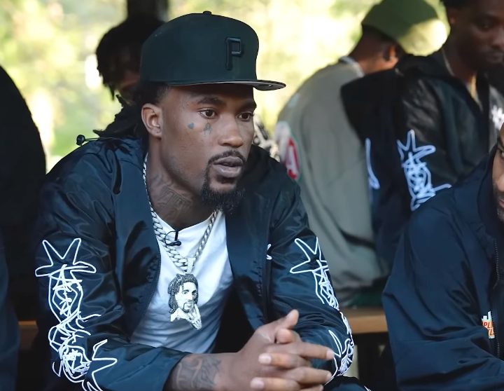 Hunxho sits talking outdoors in a black fitted cap and a black jacket with white graffiti lettering, a heavy chain and pendant at his neck.