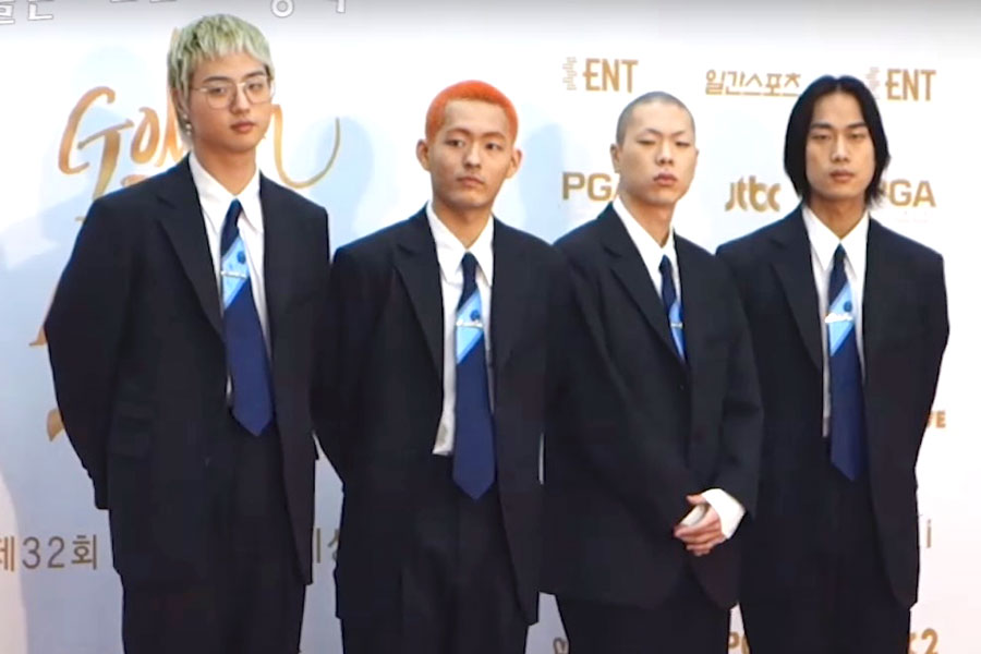 The four members of HYUKOH stand in matching dark suits and blue ties in front of an awards-show step-and-repeat banner.