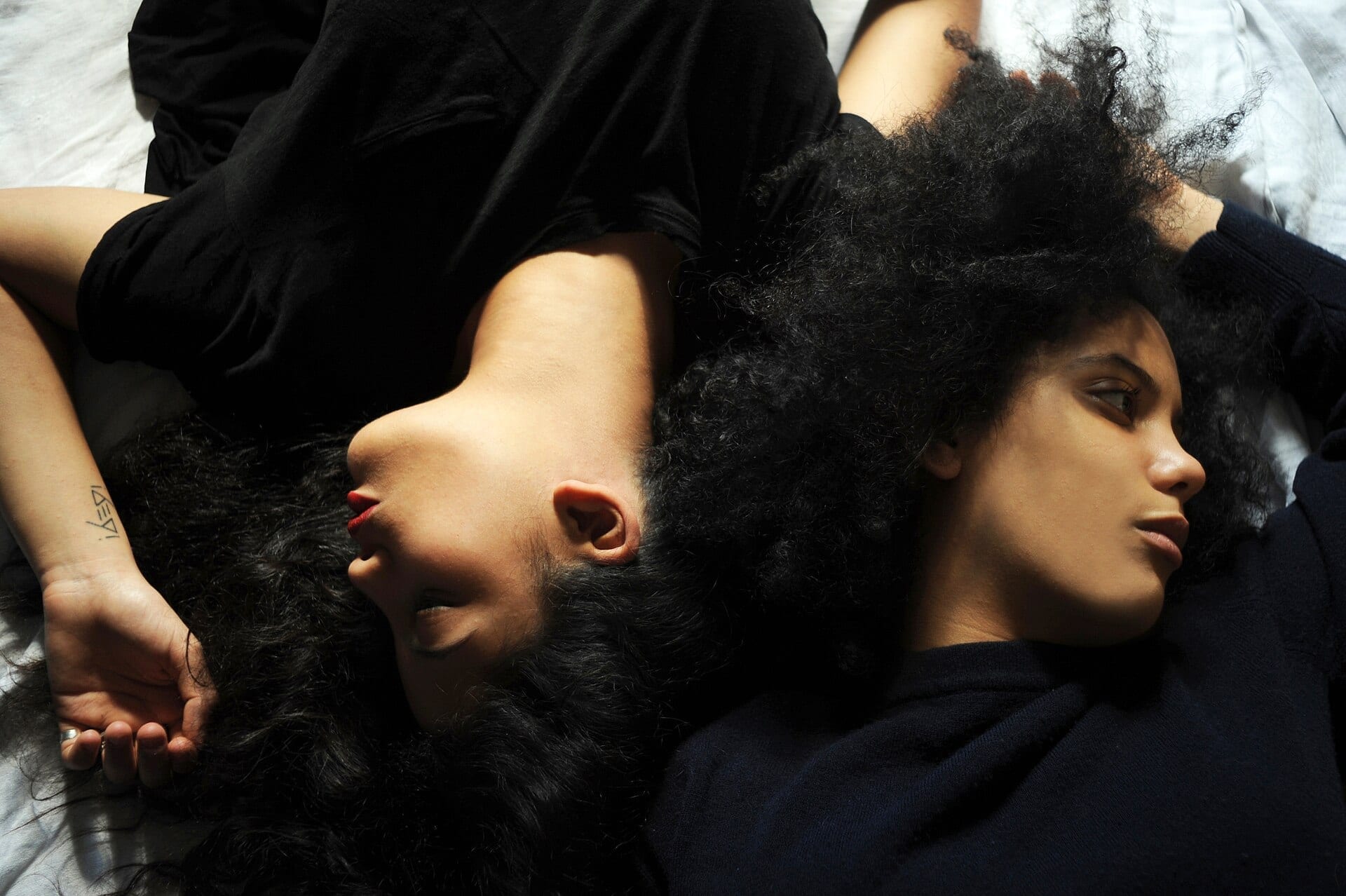 The two members of Ibeyi lie back to back with their heads touching, eyes closed, their dark curly hair spread across a white sheet.