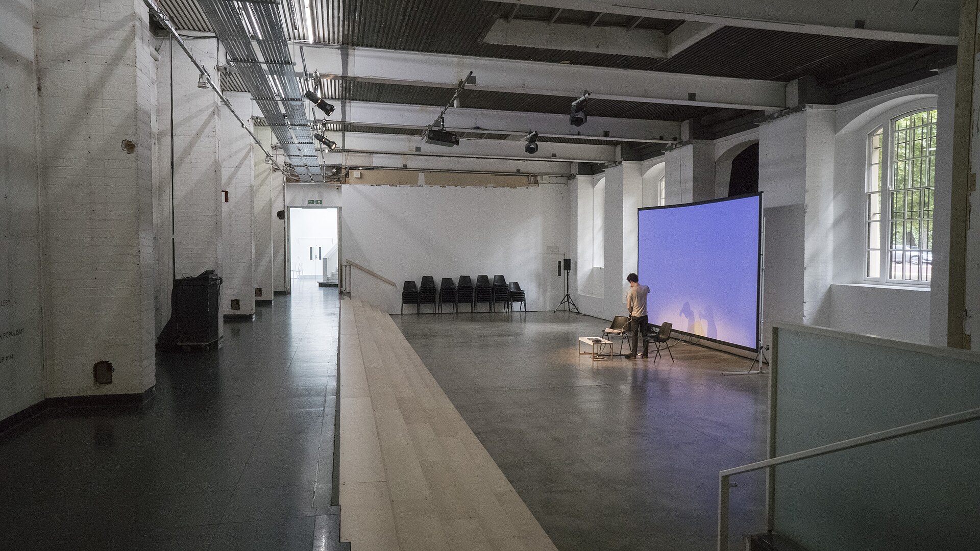 A long white-walled hall inside the Institute of Contemporary Arts, empty apart from stacked chairs and a lit projection screen at the far end.