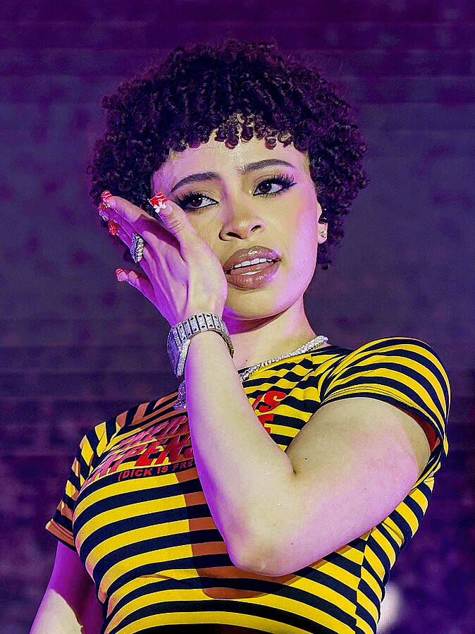Ice Spice, wearing a black-and-yellow striped top and layered jewelry, rests her hand near her cheek while performing under purple stage lighting.