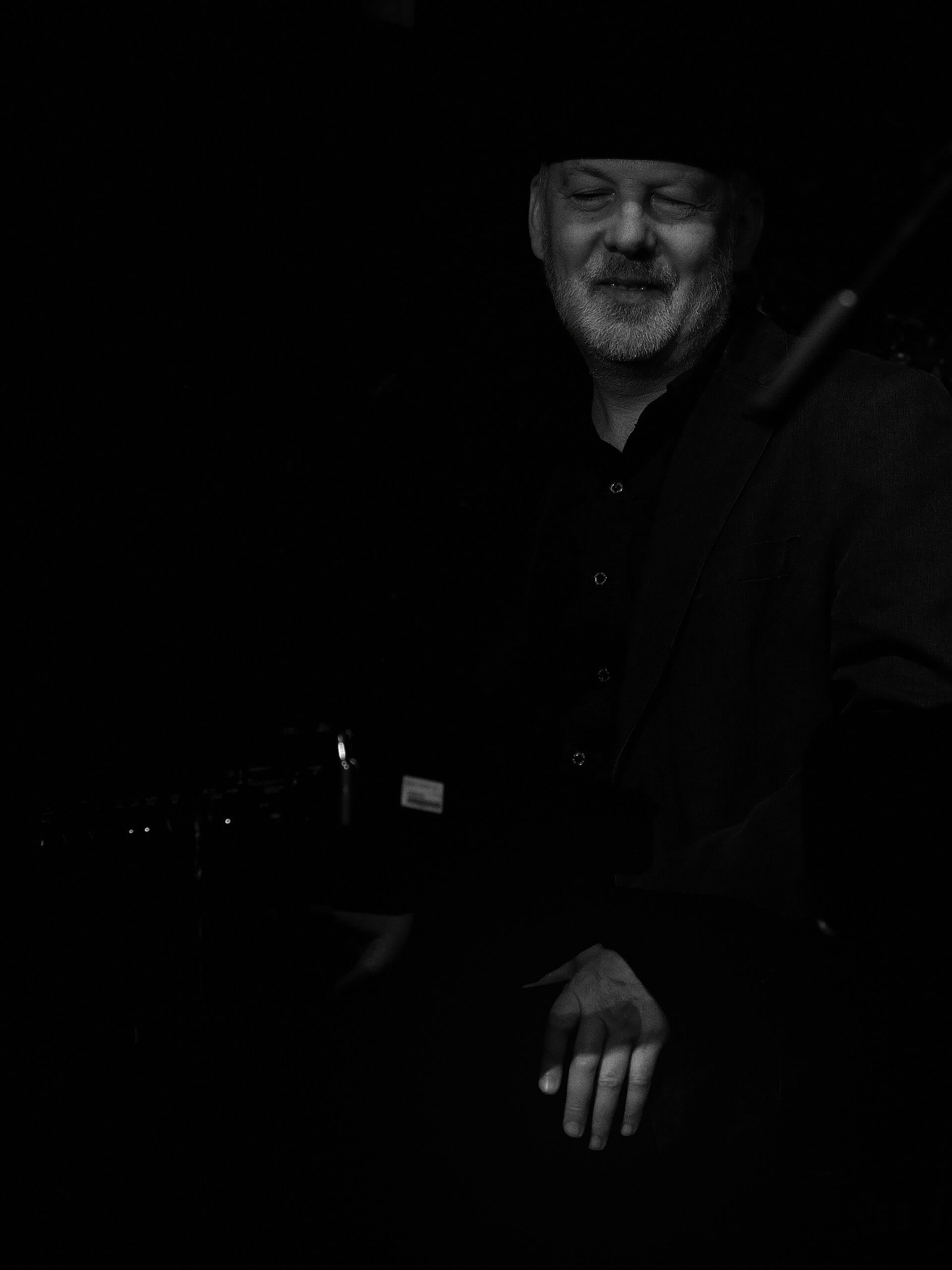 Ignasi Terraza, wearing a dark hat and shirt, plays piano with one hand extended over the keys in a moody black-and-white portrait.
