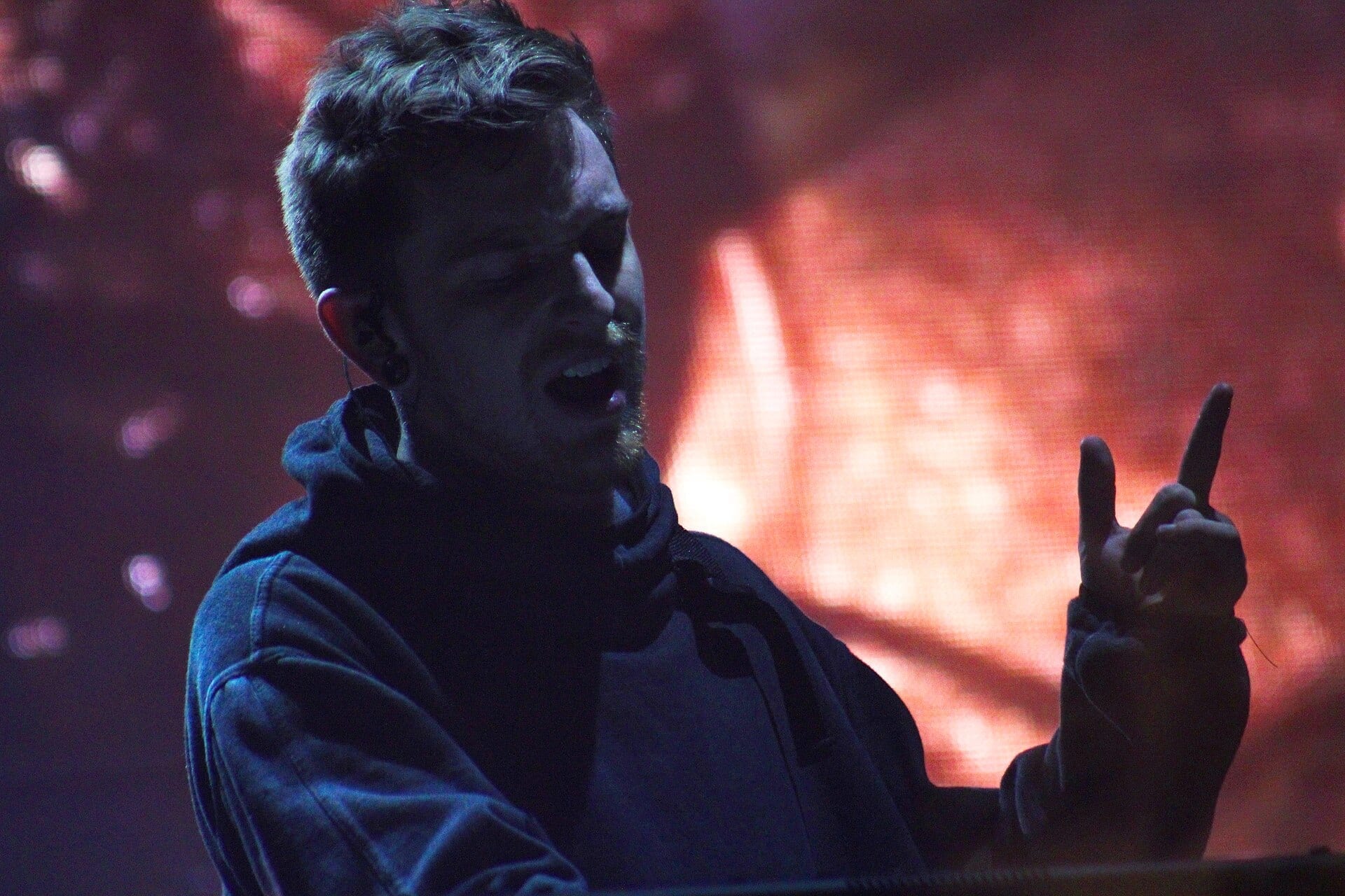 Illenium sings into a microphone with his eyes closed, wearing a hooded sweatshirt, backlit by red stage lighting and flames.