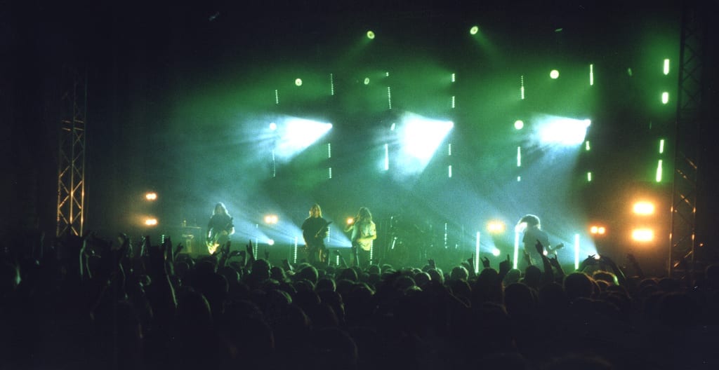 In Flames play in silhouette on a green-lit stage above a crowd of raised arms.