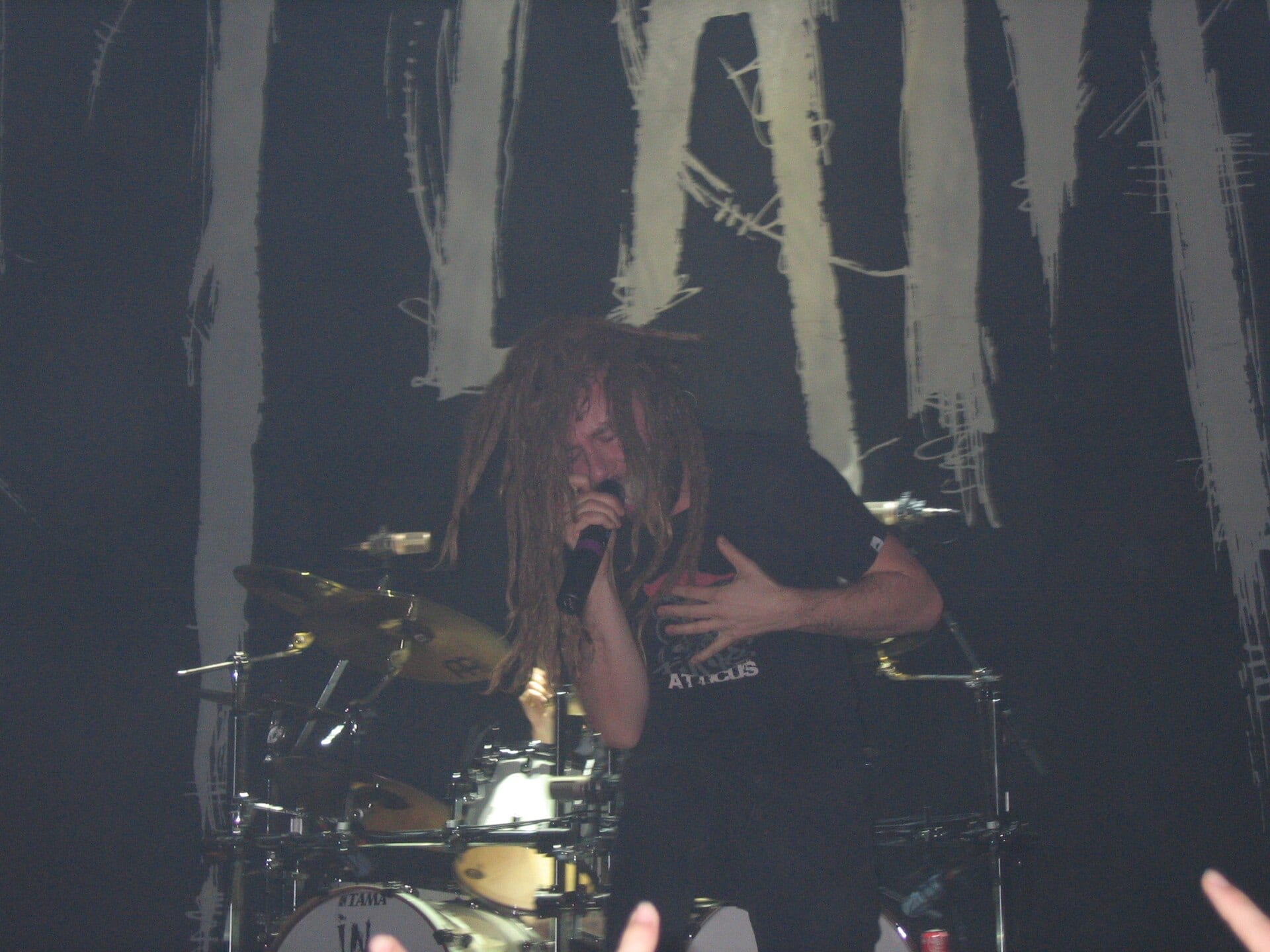 Anders Fridén sings into a handheld microphone with his long dreadlocks hanging over his face, a drum kit lit behind him.