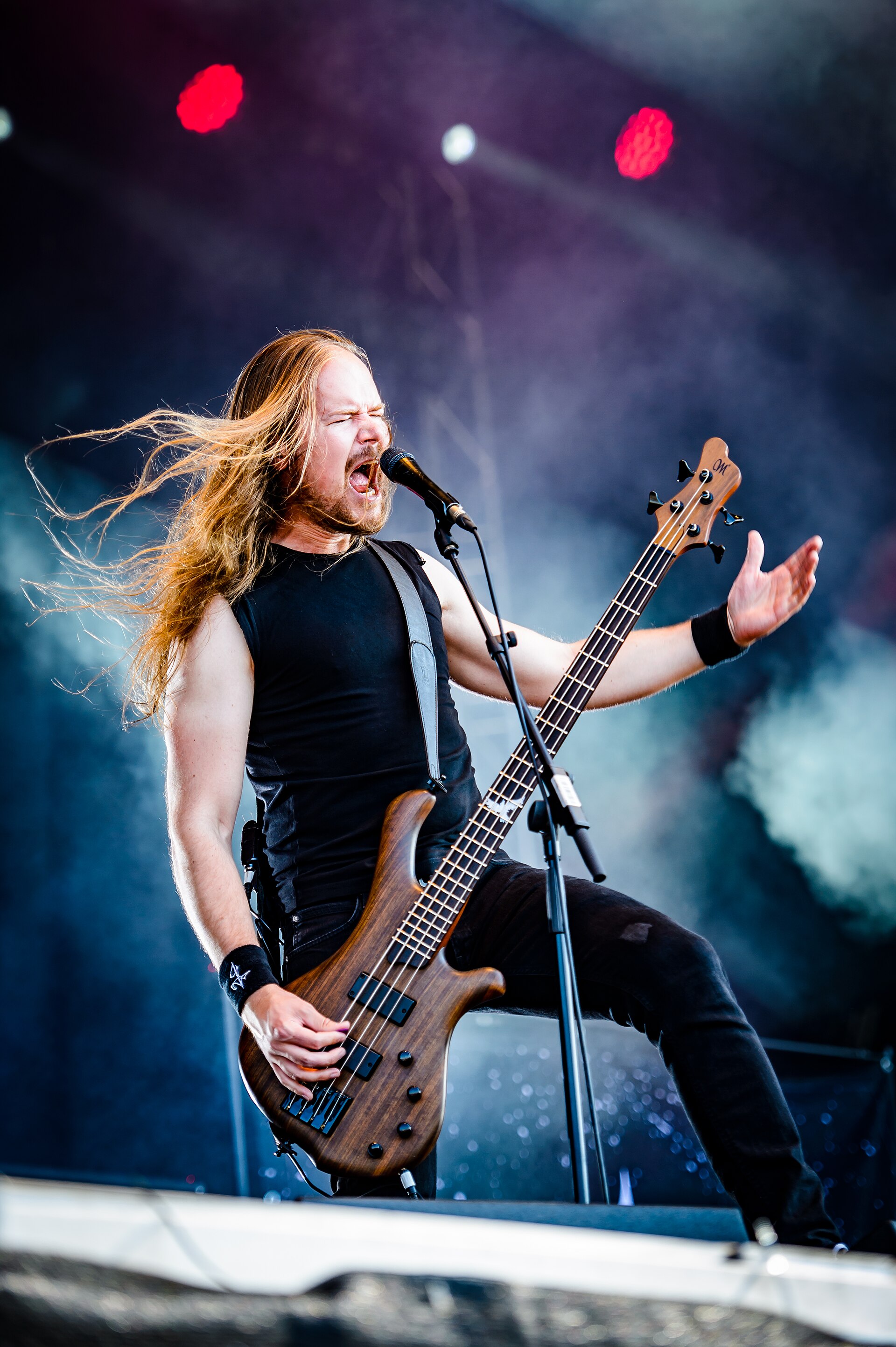 A long-haired bassist screams into a microphone mid-song, leaning back with one hand out, on a smoke-filled festival stage.