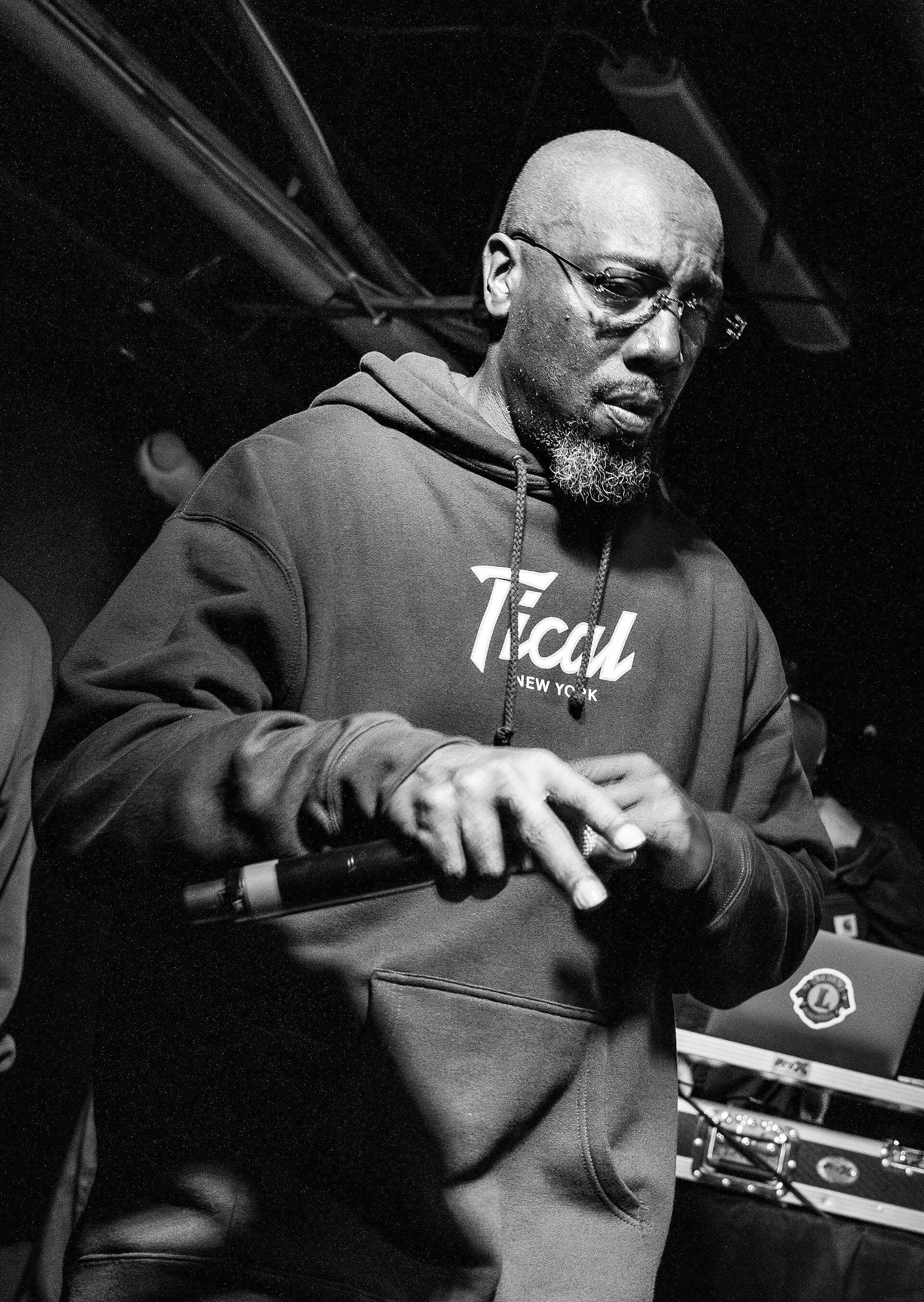 Inspectah Deck, wearing glasses and a hoodie reading Tical New York, holds a microphone while performing on stage in black and white.