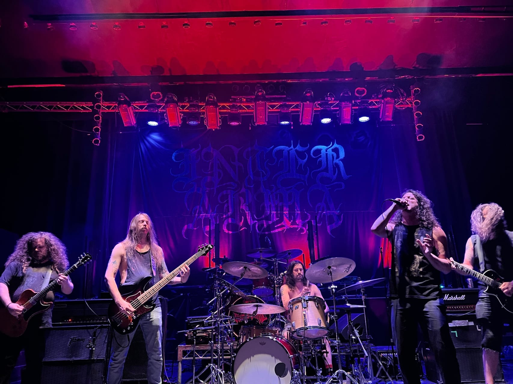 Inter Arma play a club stage in red and blue light, two guitarists and a bassist spread across the front, a drummer behind them, the band's name in blackletter on the backdrop.
