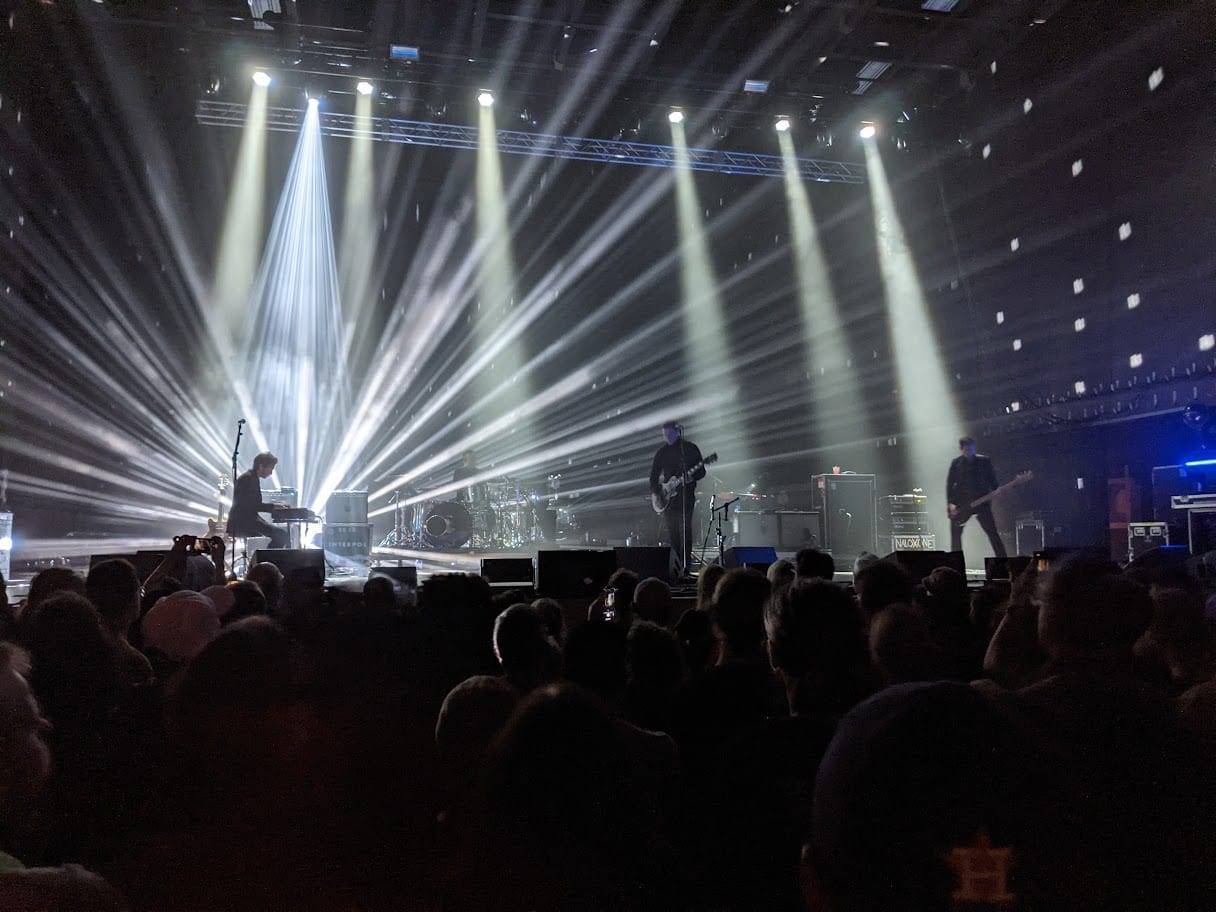 Interpol play in silhouette on a smoke-filled stage lit by fanned white spotlights, with a crowd watching from the foreground.
