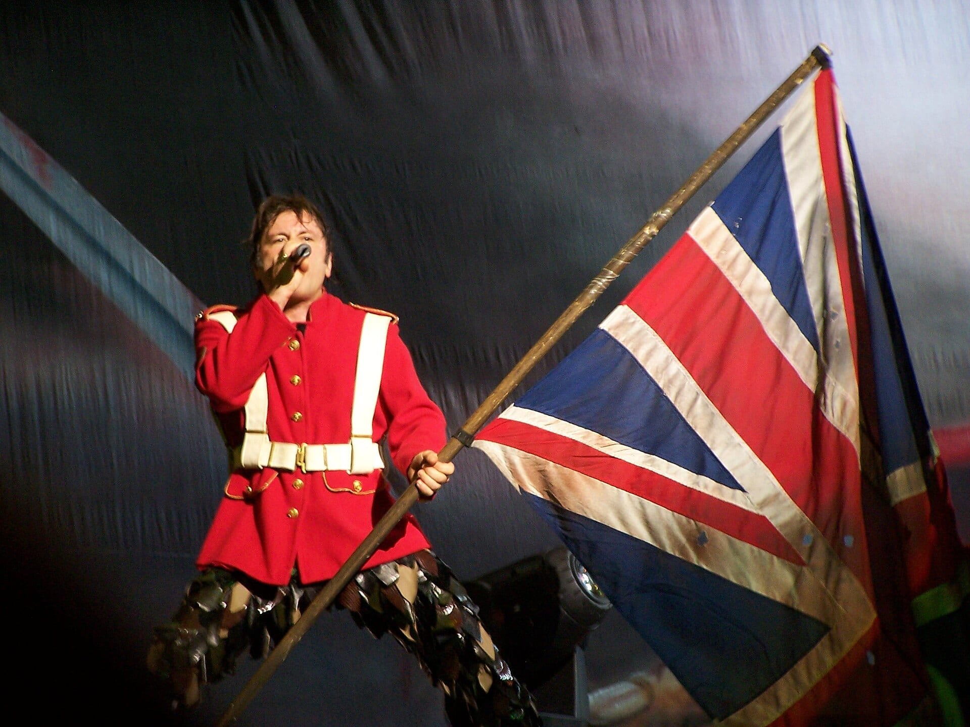Bruce Dickinson sings into a microphone in a red military tunic while holding a Union Jack on a wooden pole.