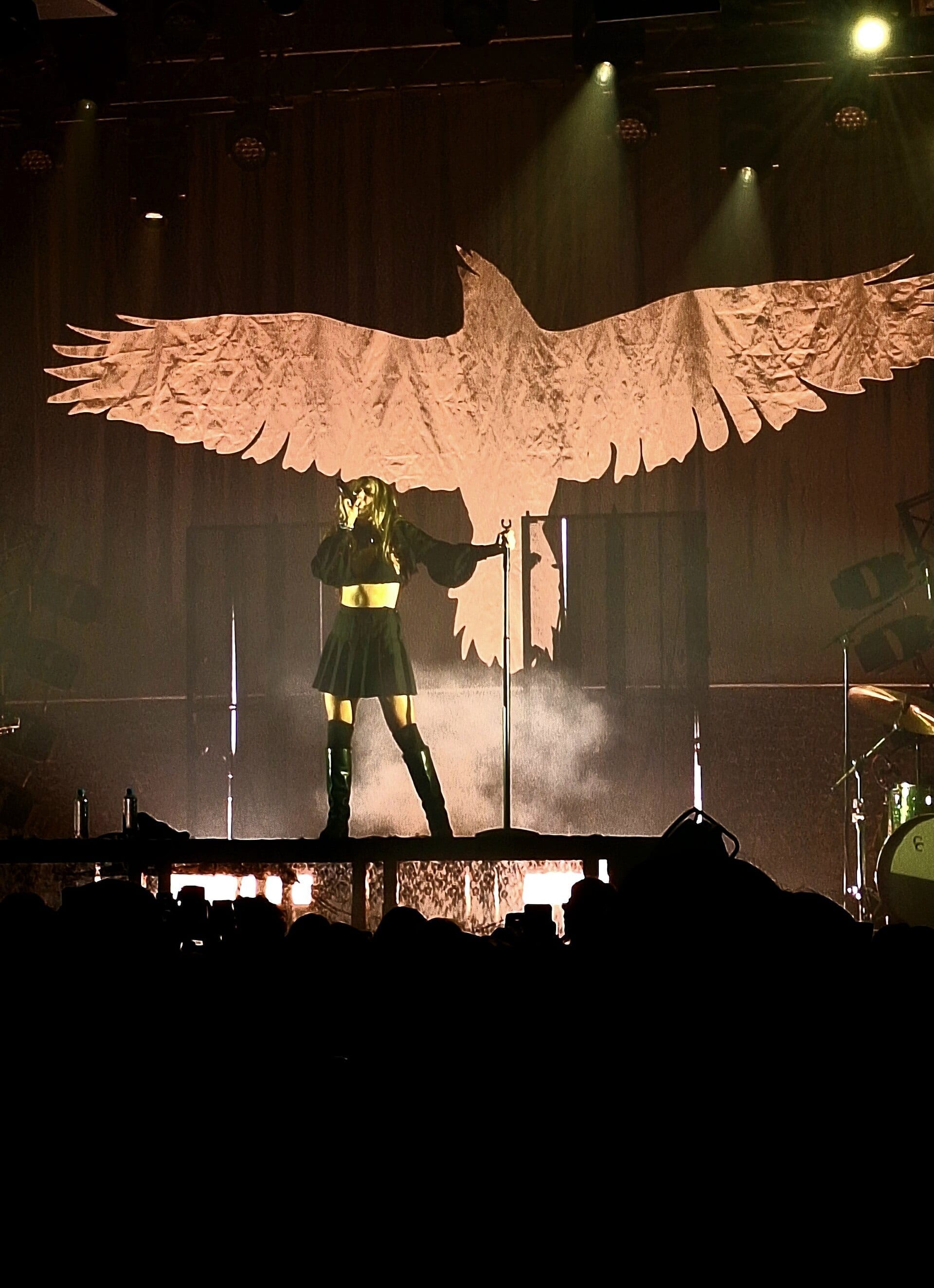 Isabel LaRosa stands silhouetted on a lit stage in a dark skirt and knee-high boots, holding a microphone stand, beneath a giant projected outline of a bird with outstretched wings.