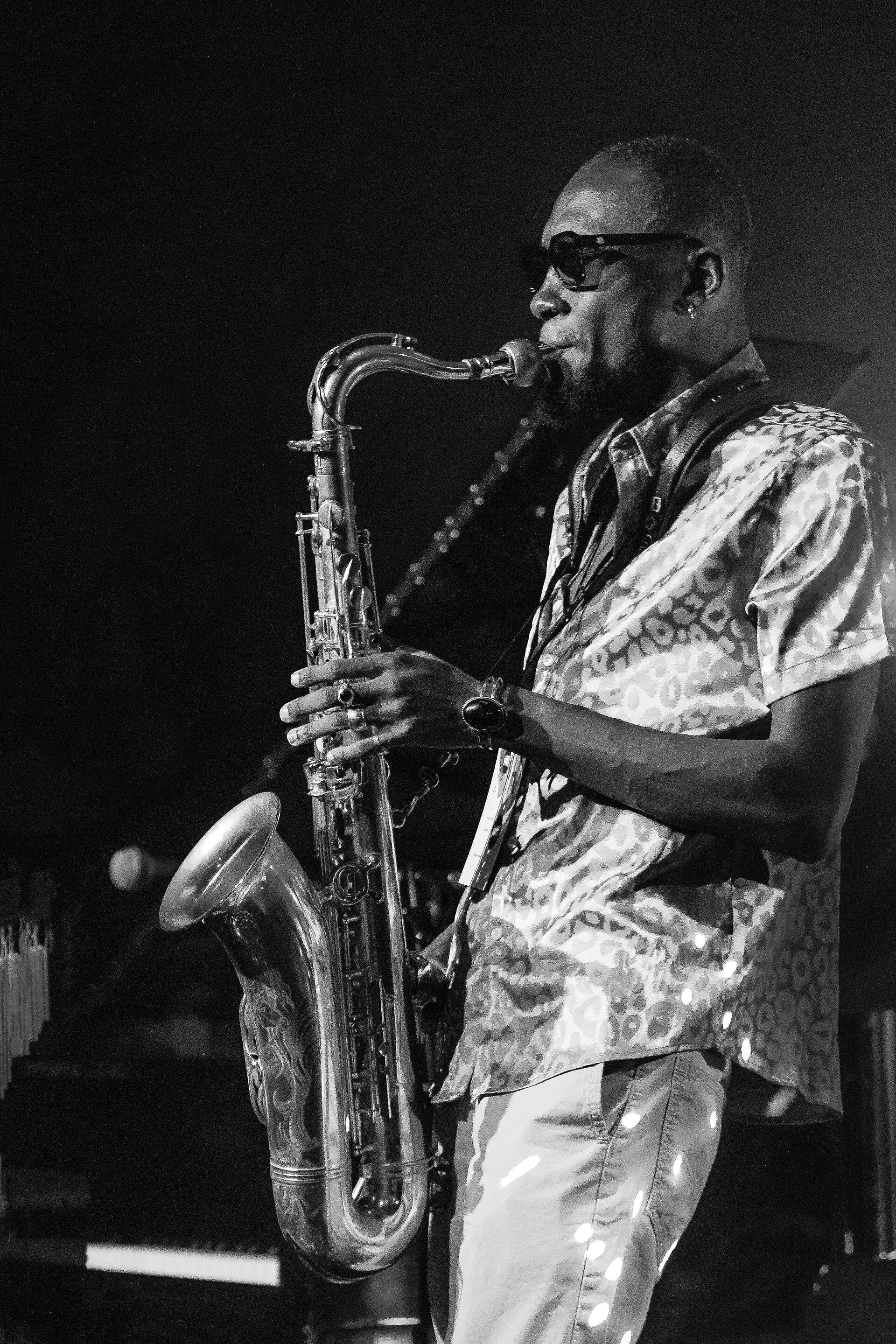 Isaiah Collier plays saxophone on stage, wearing sunglasses and a patterned shirt, in a black and white photo.