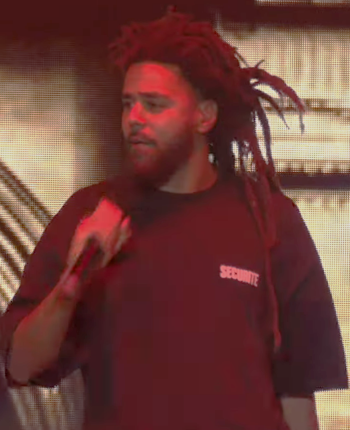 J. Cole raps into a microphone in a red-lit stage shot, wearing a maroon T-shirt with his locs pulled back.