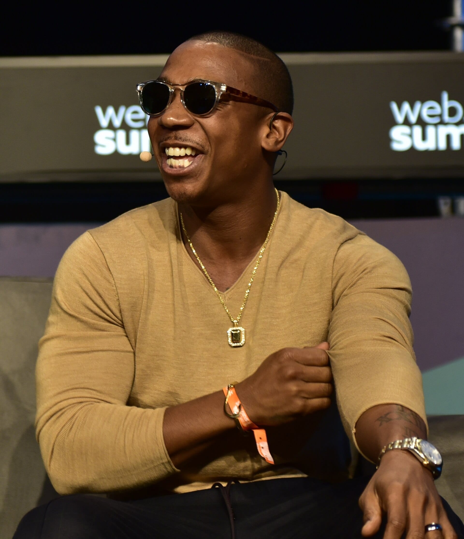 Ja Rule laughs while wearing sunglasses and a headset microphone, seated on stage in a tan sweater with a gold chain and pendant.
